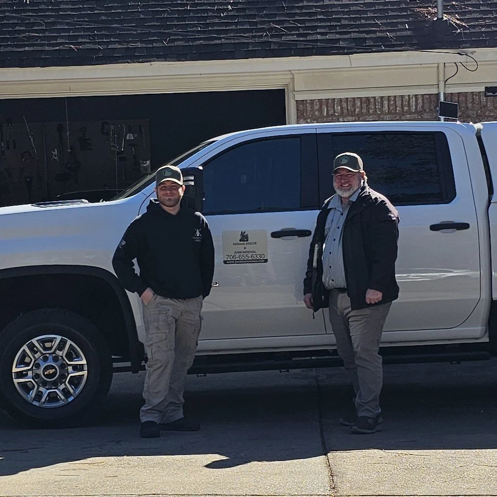 Two men standing in front of a white vehicle with a garage and tools in the background. Both men wear caps and outdoor clothing, smiling at the camera.