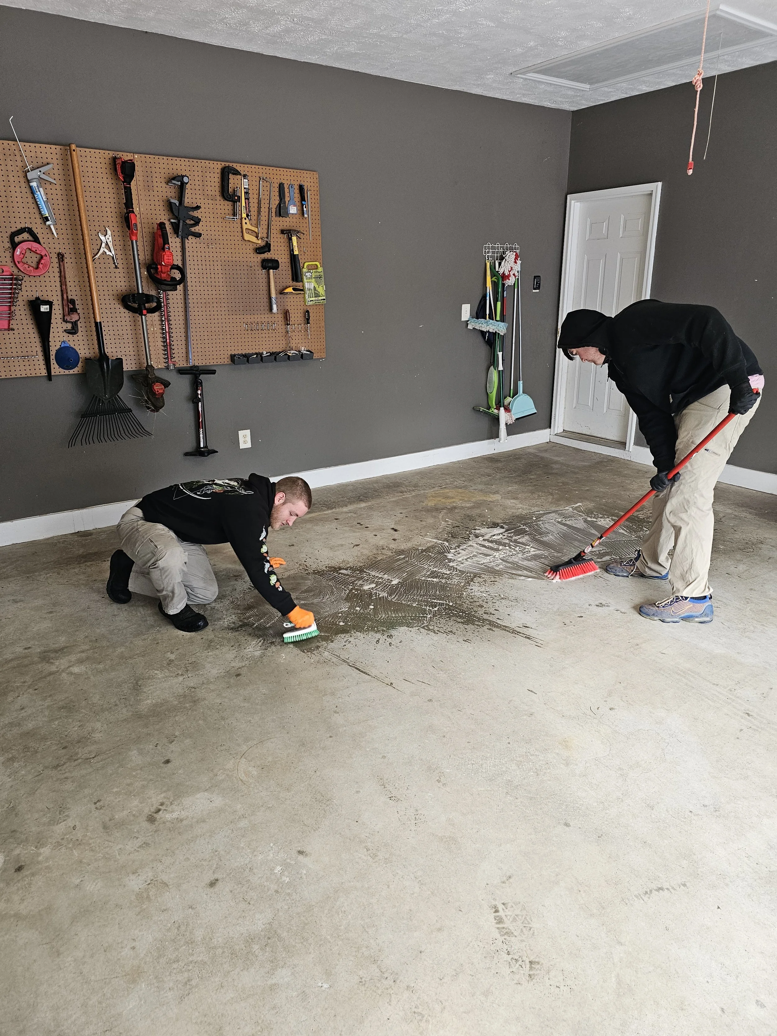 Two men are working on the floor of a garage, cleaning or preparing the concrete surface. One is crouching and the other is standing with a broom. The garage has a pegboard on the wall holding various tools, cleaning supplies, and equipment.