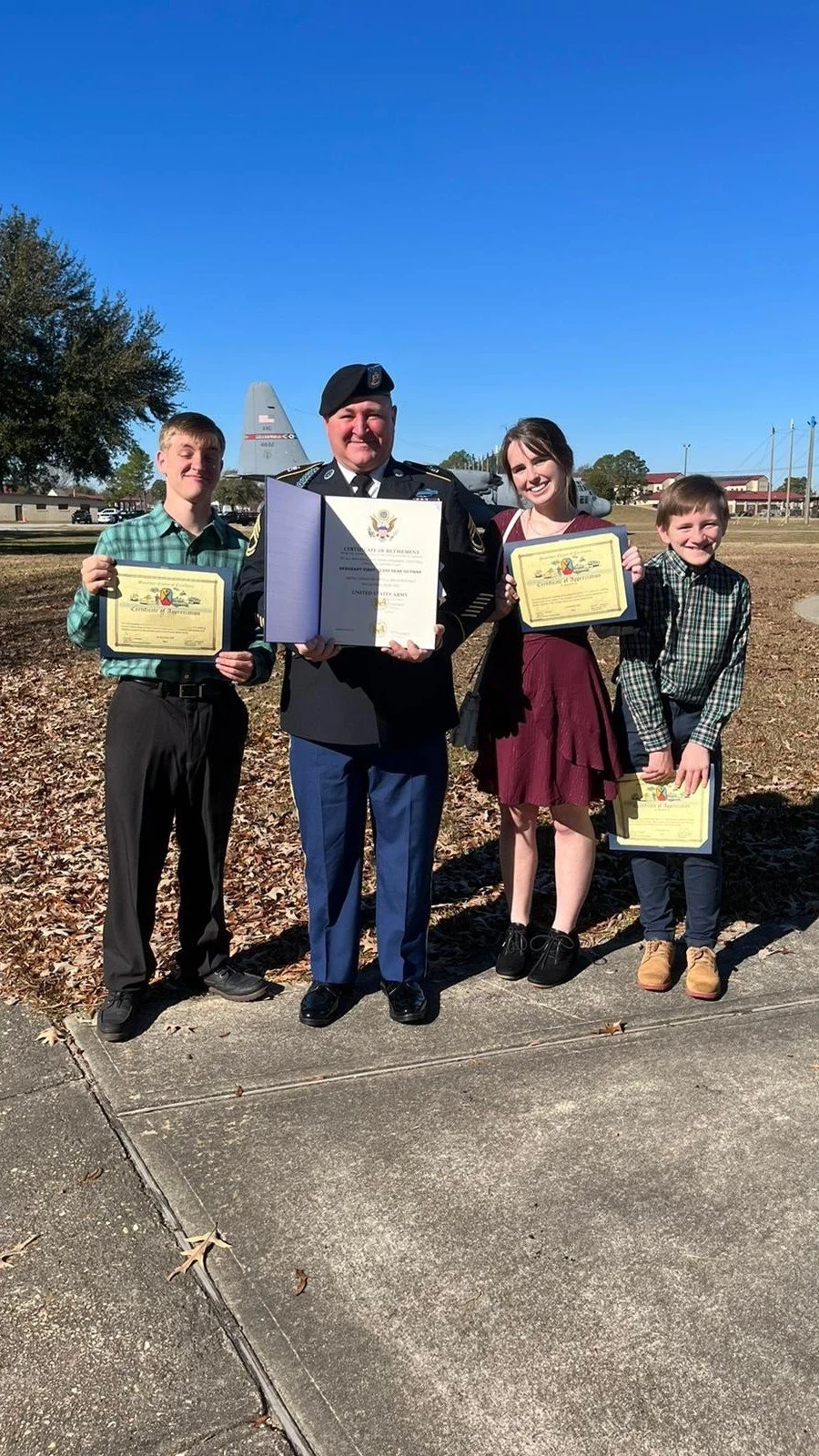 A group of four people, including an Army officer and three children, standing outdoors on a sunny day with an aircraft in the background. The children are holding certificates, and the officer is displaying a certificate folder, celebrating an achievement or award event.