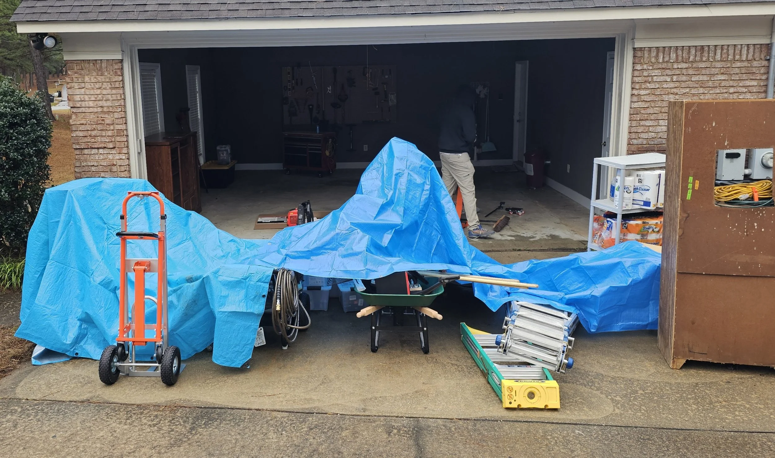 Garage under renovation with tools and equipment, covered with blue tarp, including a hand truck, extension cords, ladder, and a toolbox, with a person working inside.