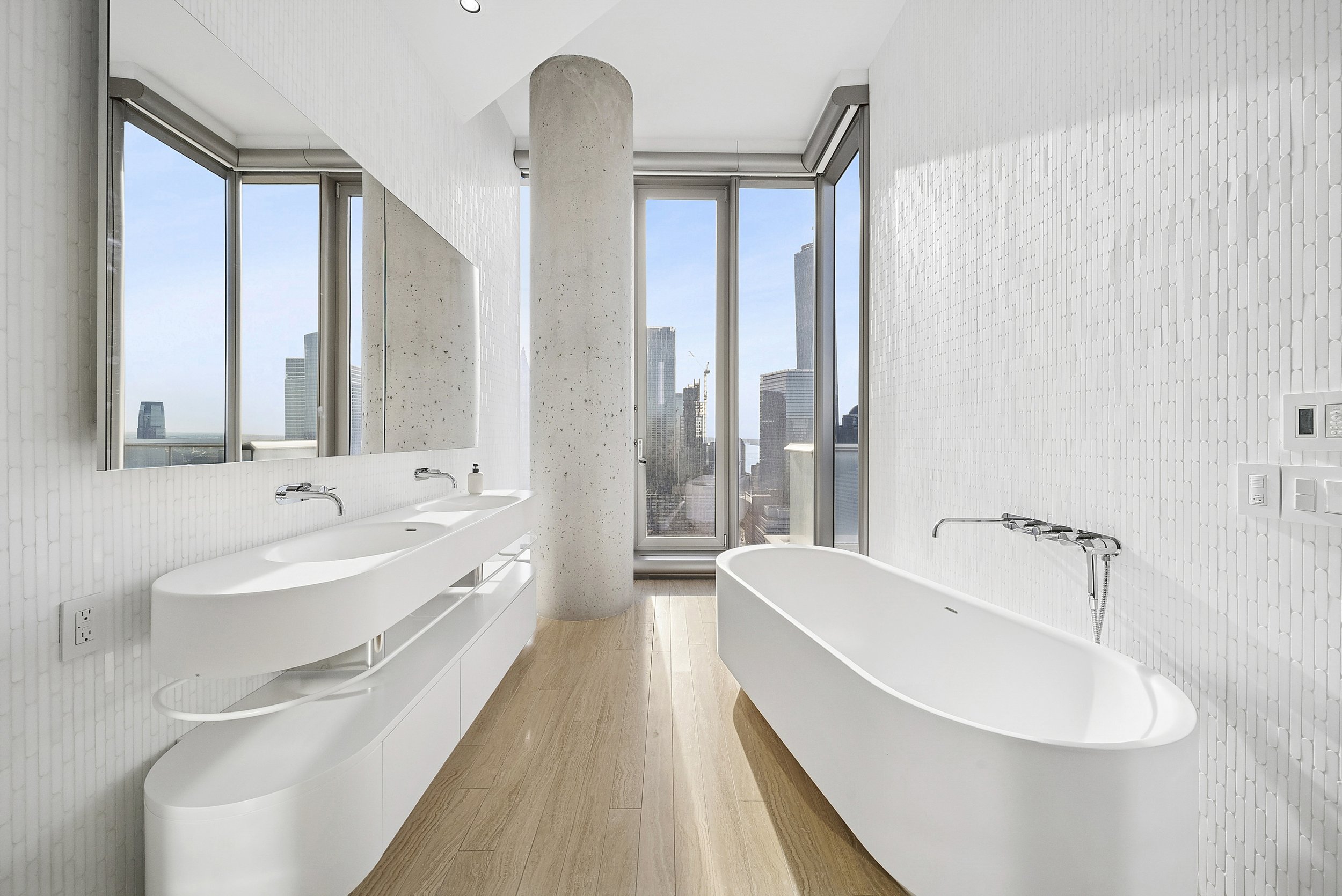 Modern white bathroom with freestanding bathtub, dual sinks, large windows with city skyline view, and wood flooring.