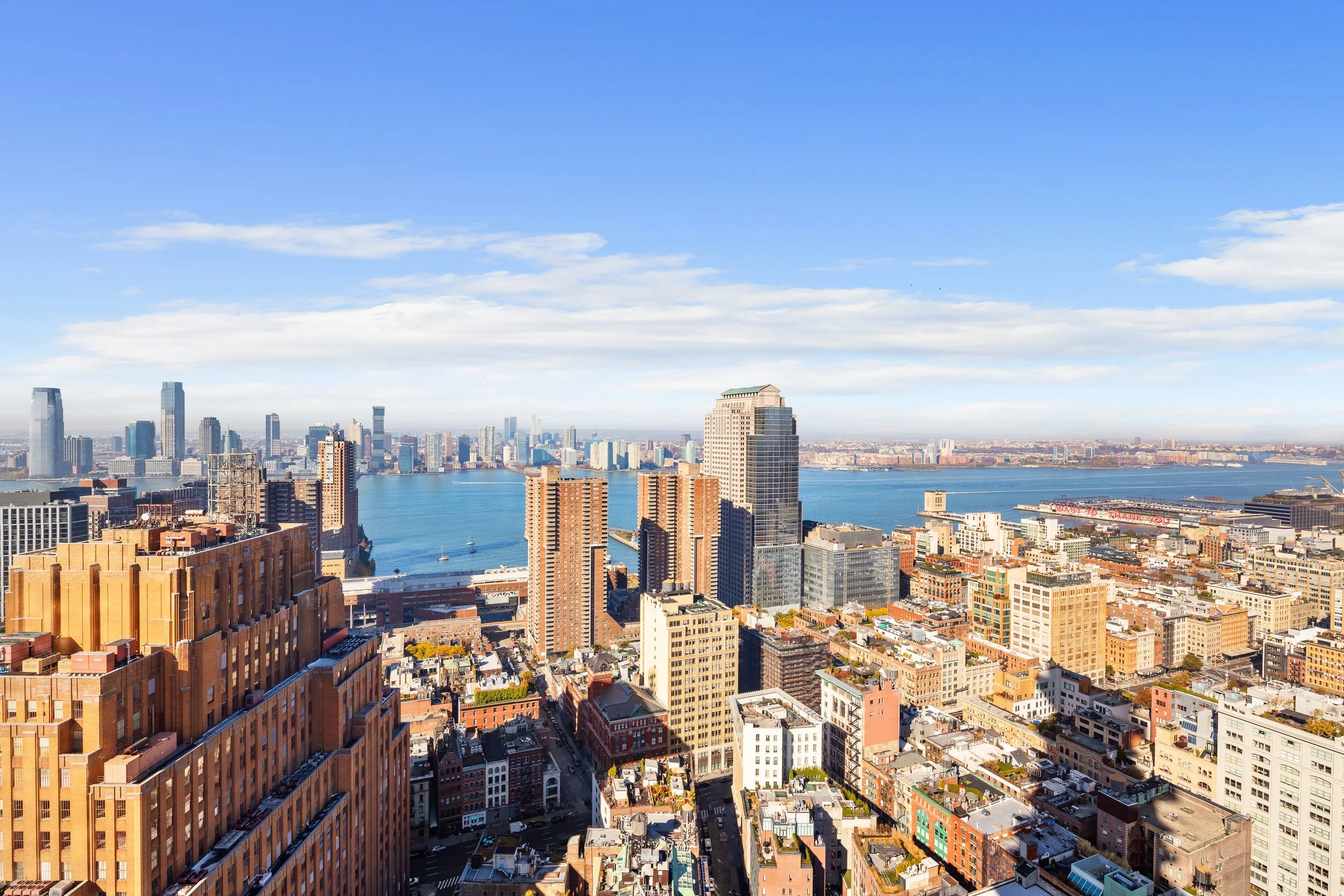 A city skyline with tall buildings and a river in the background under a blue sky with scattered clouds.
