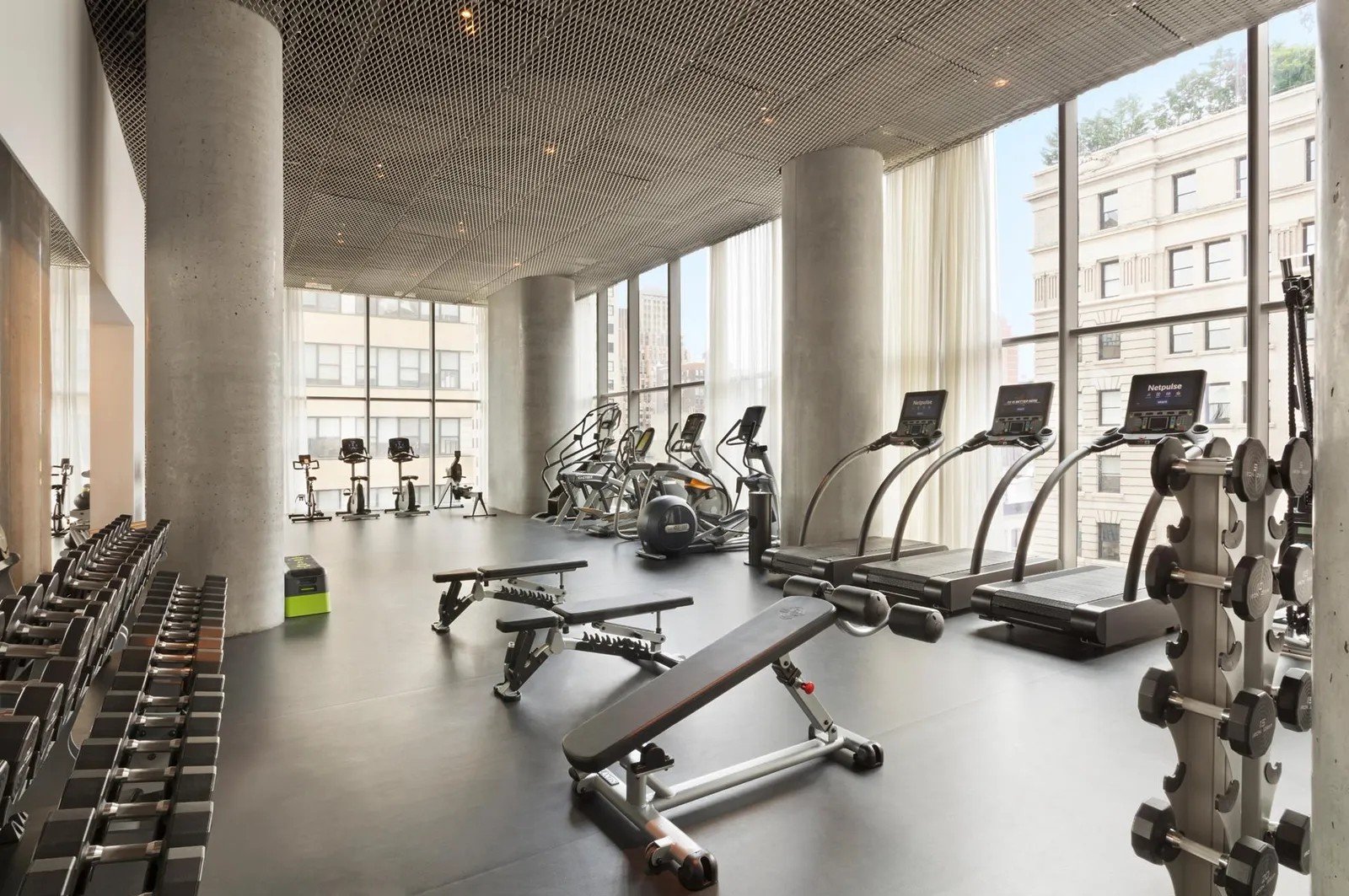 Modern gym with cardio equipment, weight racks, and a bench near large windows with city view.