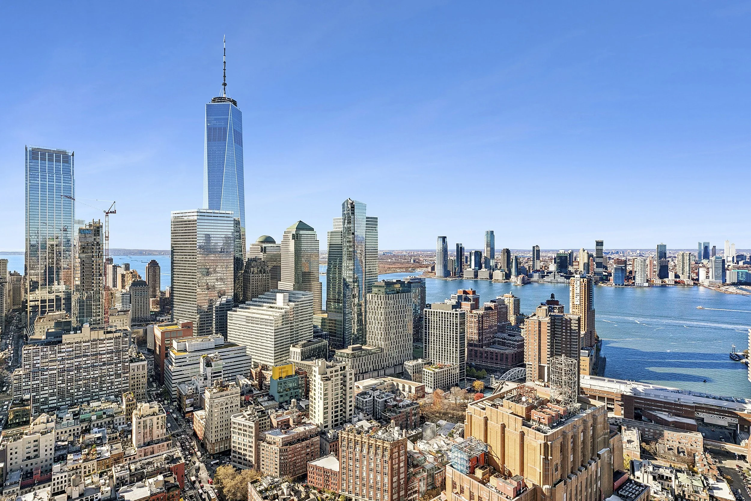 A city skyline featuring One World Trade Center in New York City with tall skyscrapers and the Hudson River visible on a clear, sunny day.