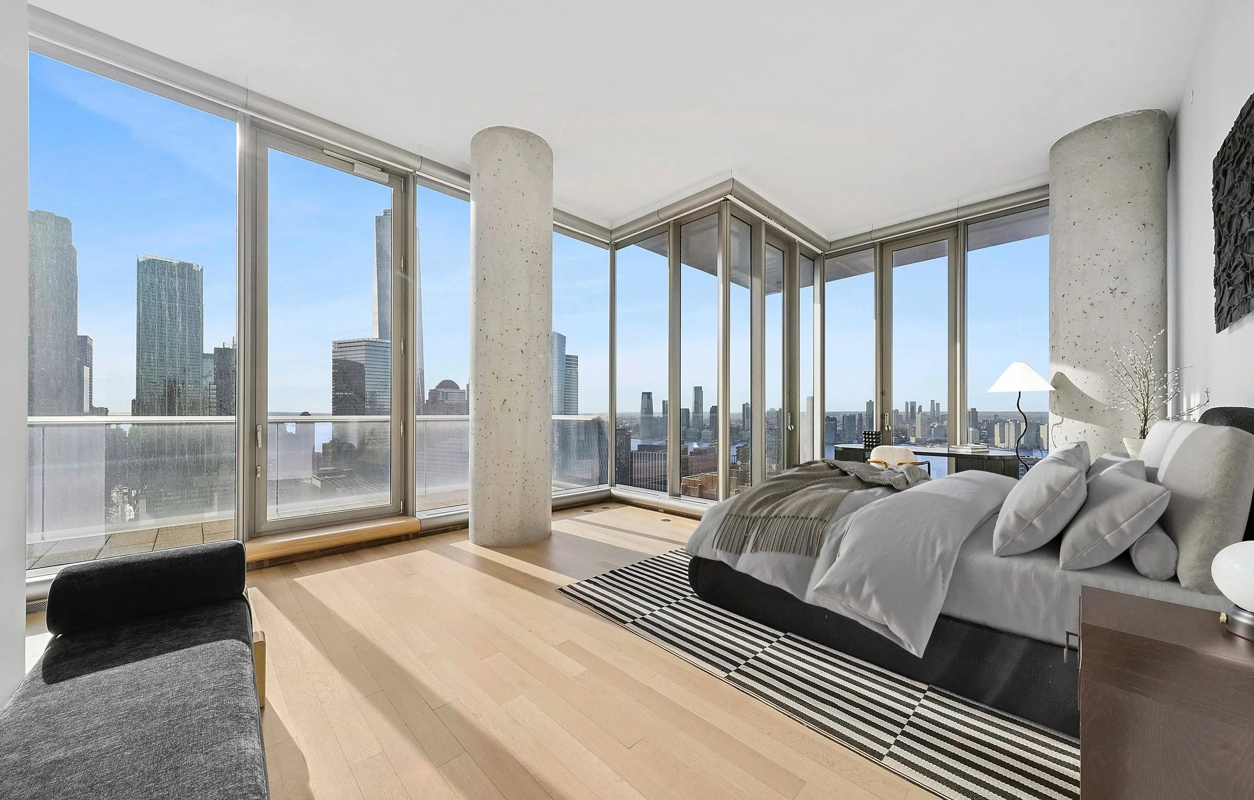 Modern bedroom with large floor-to-ceiling windows offering city skyline views, a bed with white bedding and pillows, a black and white striped rug, a bedside table with a lamp and decorative branches, and light wooden flooring.