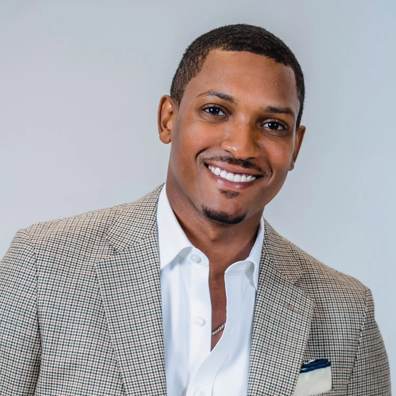 Portrait of a smiling man wearing a white shirt and a checked blazer, with a light background.