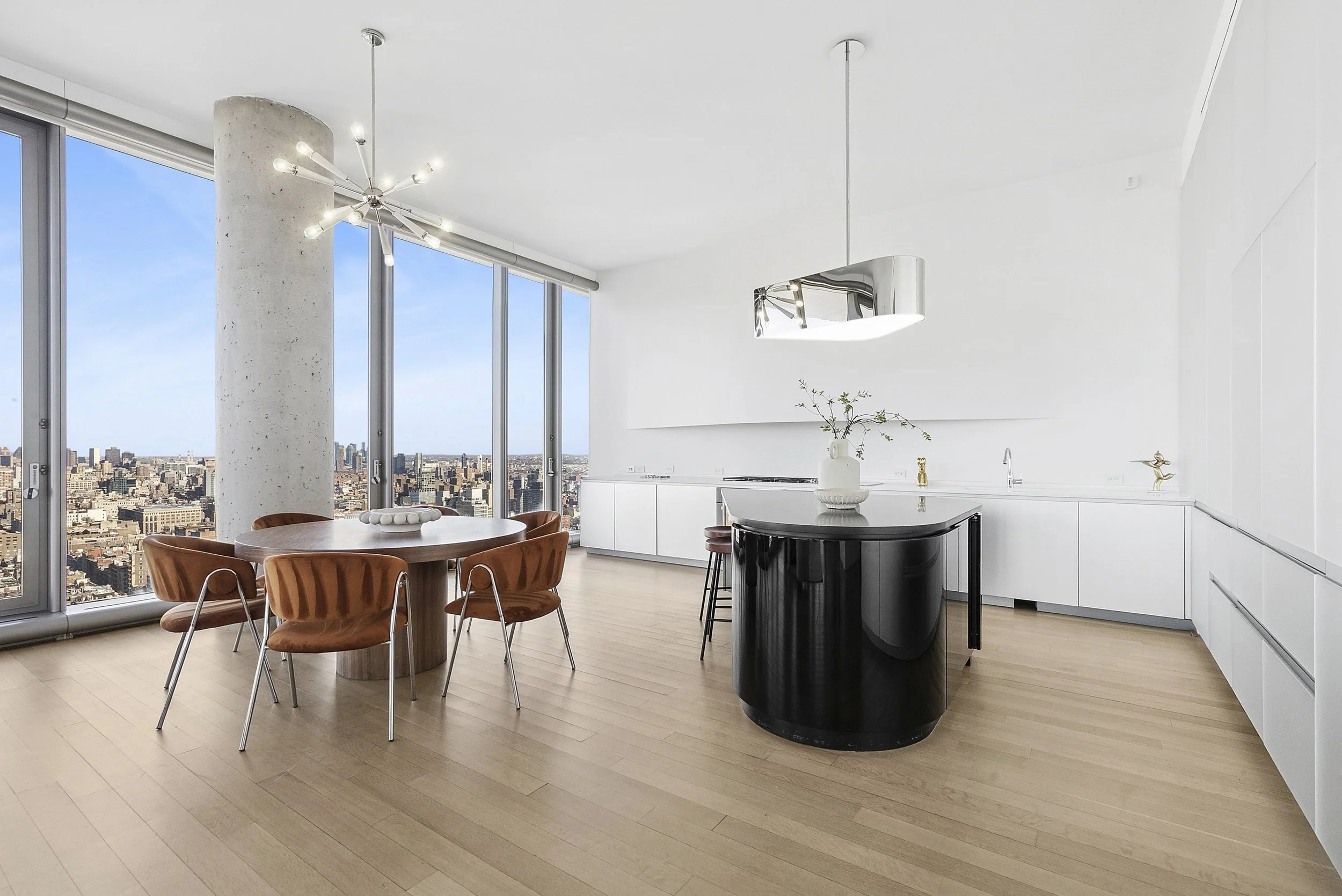 Modern high-rise kitchen with light wood flooring, white cabinets, black curved kitchen island, and floor-to-ceiling windows showcasing a cityscape.