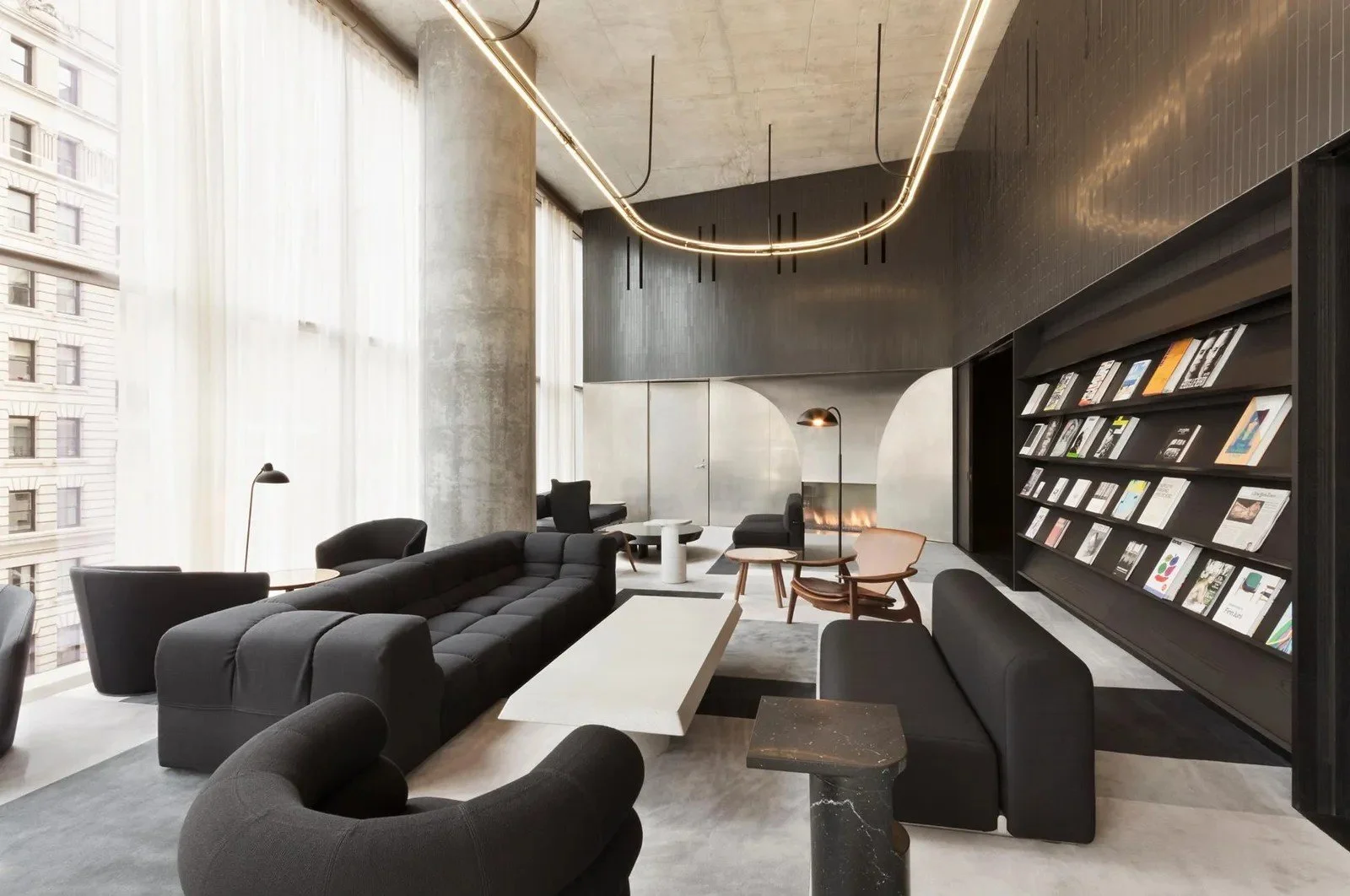 Modern lobby with black couches, armchairs, a white coffee table, large windows with sheer white curtains, bookshelves, and ceiling lighting.