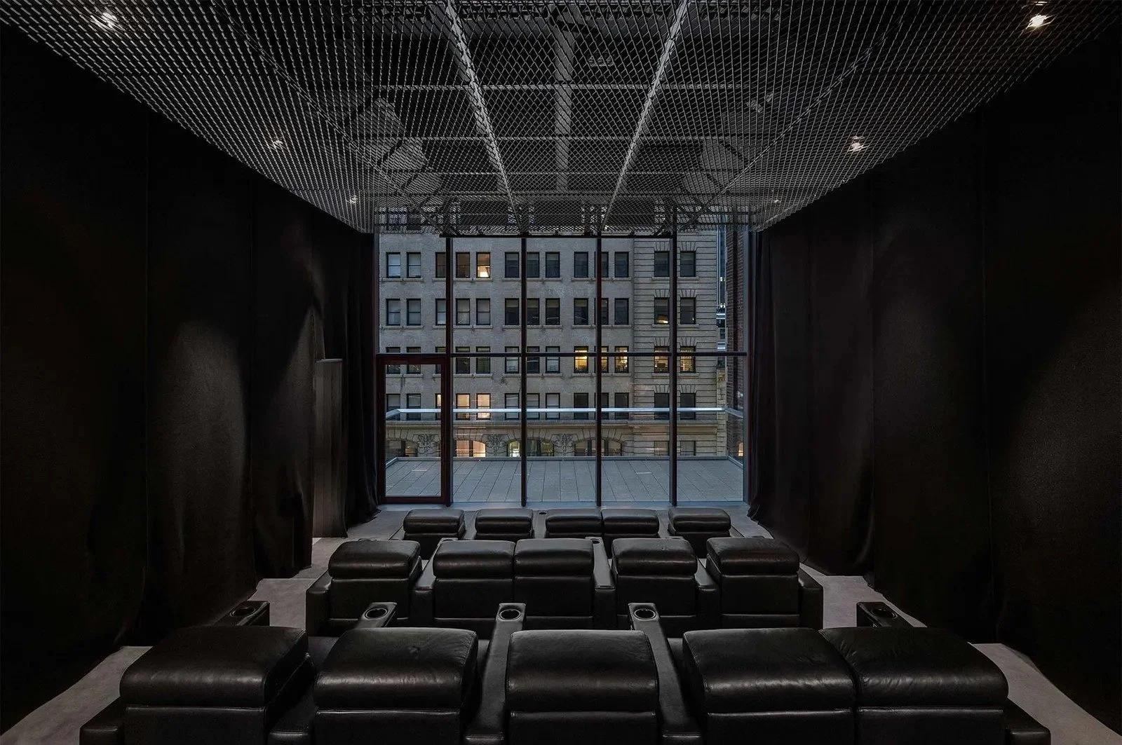 Interior of an empty black theater or screening room with leather seats facing a large window showing neighboring buildings.