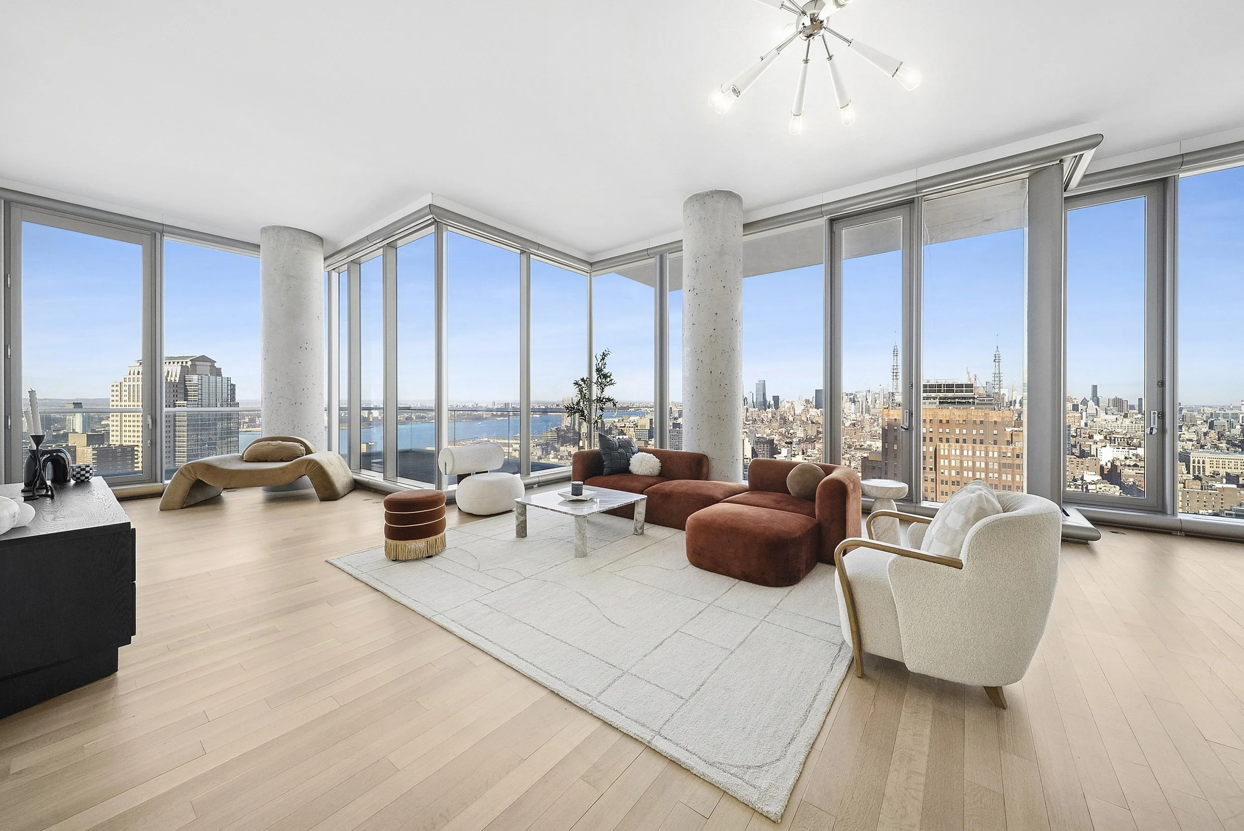 Luxury high-rise living room with panoramic city views, featuring large windows, a modern chandelier, a beige chaise lounge, a white ottoman, a rust-colored sectional sofa, a white armchair, a marble coffee table, and minimalistic decor.