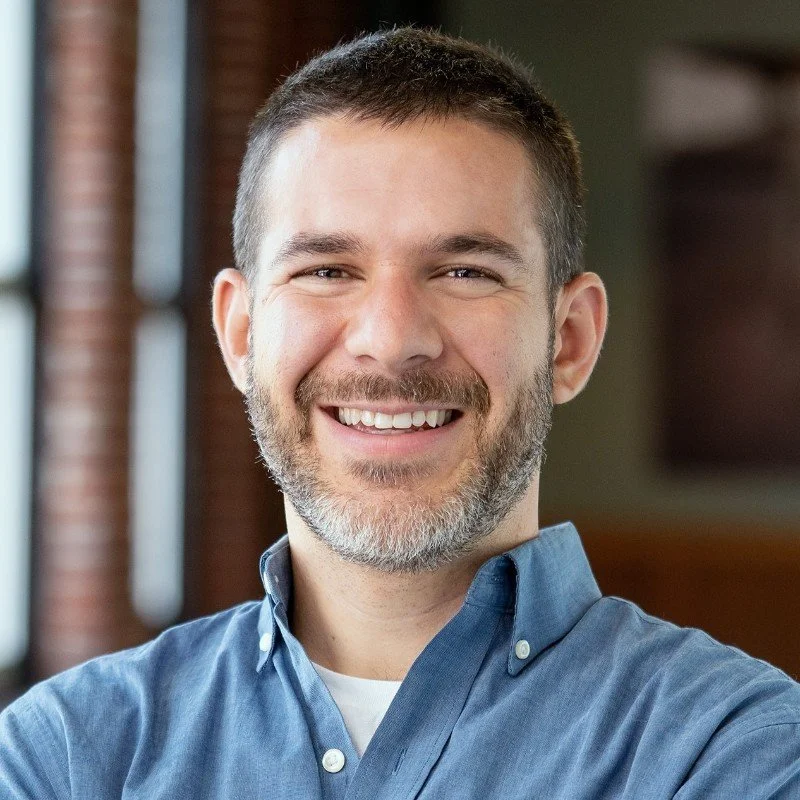 A smiling man with short brown hair and a graying beard wearing a blue button-up shirt.