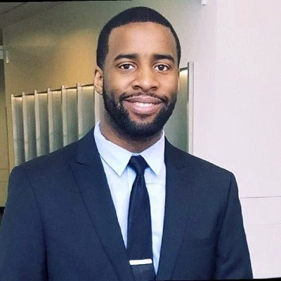 A man in a navy blue suit and tie smiling, indoor location with a partition behind him.