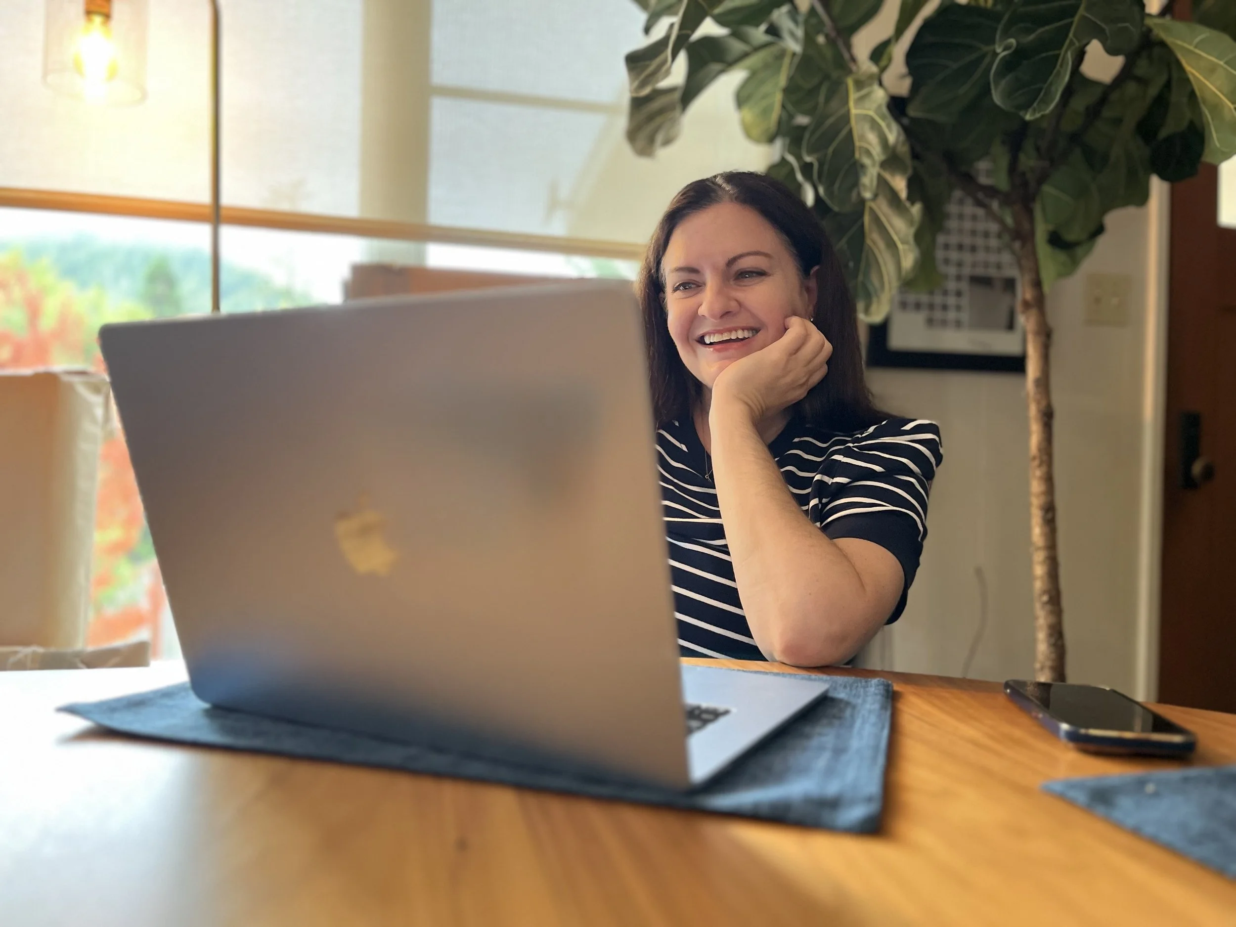 Woman with dark hair smiling and looking at a silver Apple MacBook laptop on a wooden table, with a large green plant and window in the background.