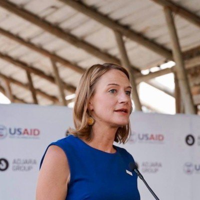 Woman in blue dress speaking at a podium with USAID and other logos in the background.