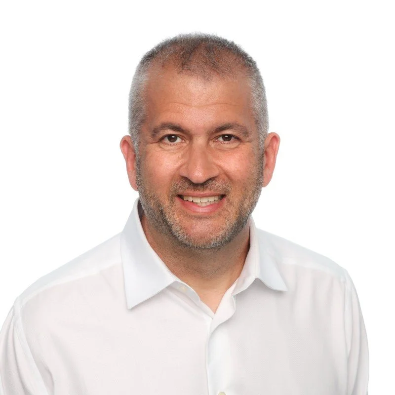 A smiling middle-aged man with short gray hair and a beard, dressed in a white collared shirt, against a plain white background.