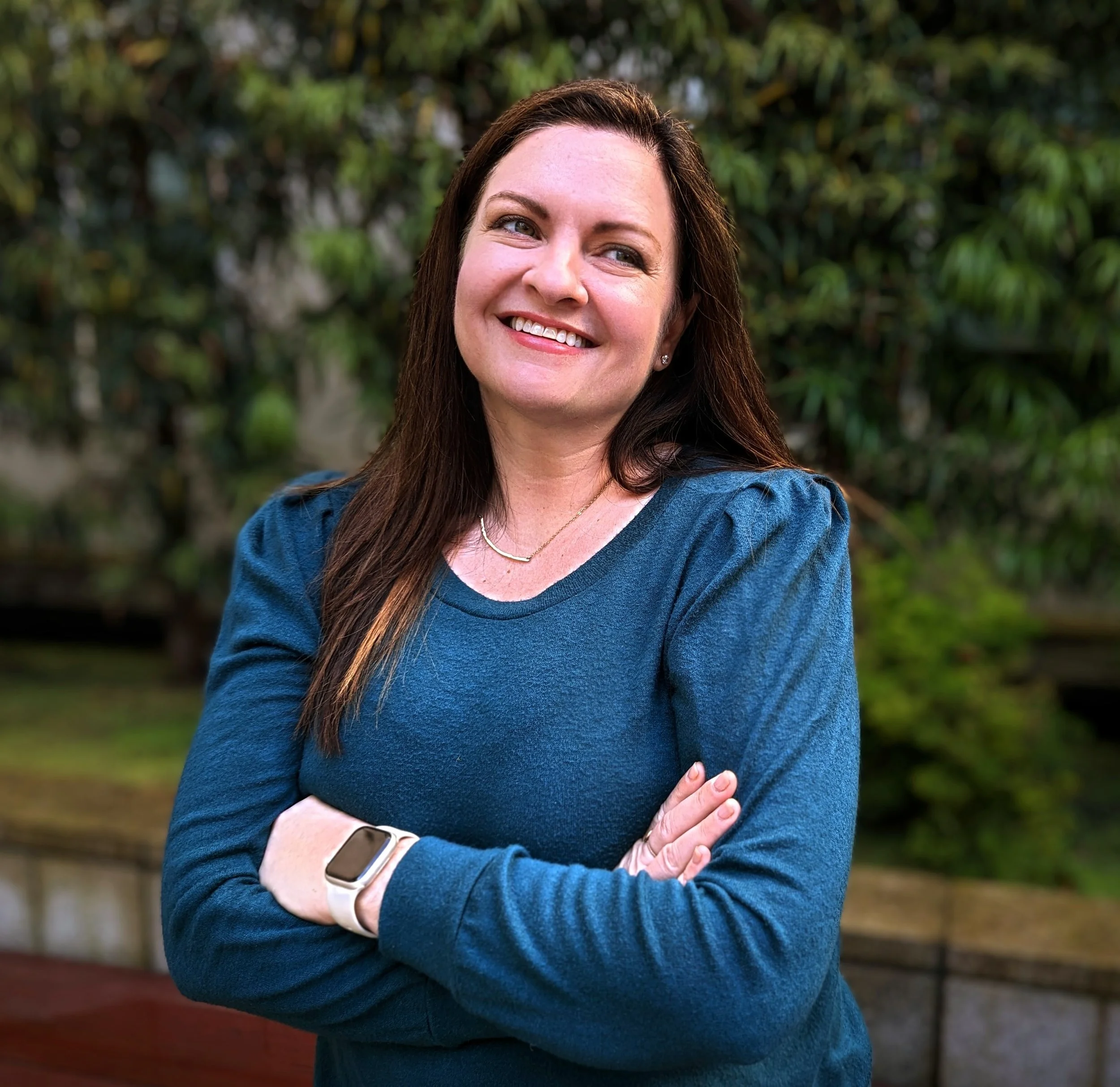 Smiling woman with brown hair standing outdoors with arms crossed, wearing a blue long sleeve shirt and a smartwatch.
