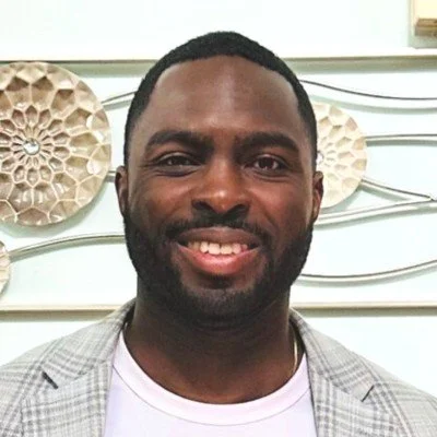 Portrait of a smiling man with short dark hair and a beard, wearing a light gray checkered blazer over a white shirt, standing in front of a light-colored wall with decorative round wall hangings.