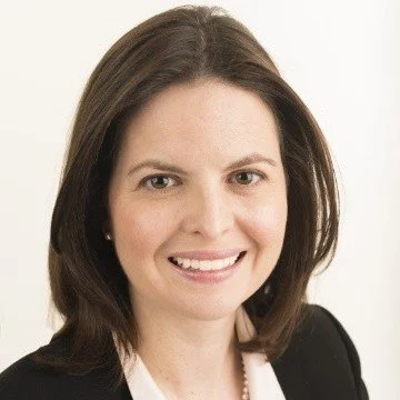 Headshot of a woman with shoulder-length brown hair, smiling, wearing a black blazer and white top against a white background.