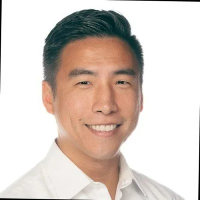 Portrait of a smiling man with short dark hair wearing a white collared shirt against a white background.