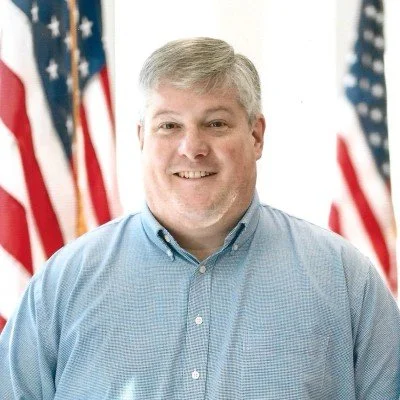 A man smiling in front of American flags, wearing a light blue button-up shirt.