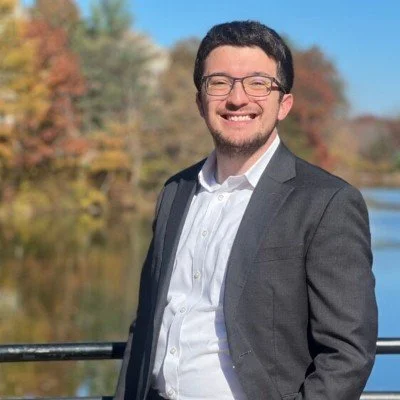A smiling man wearing glasses and a gray suit jacket outdoors with a river and autumn-colored trees in the background.