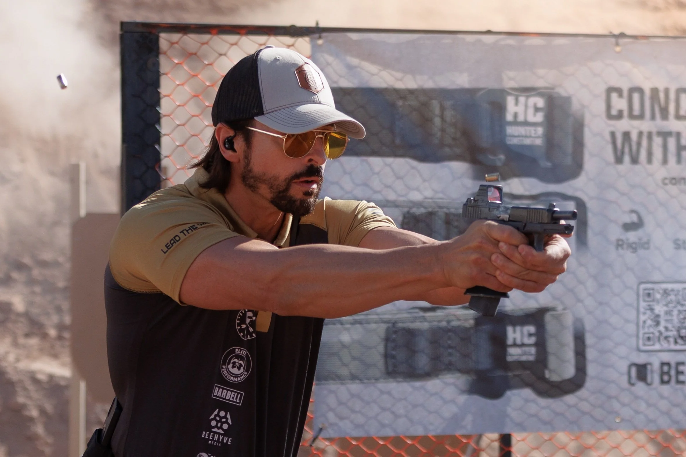 A man wearing sunglasses and tactical gear aiming a handgun at an outdoor shooting range with barrels and a mesh cover in the background. One of Arsenals Influencers/ Creators.