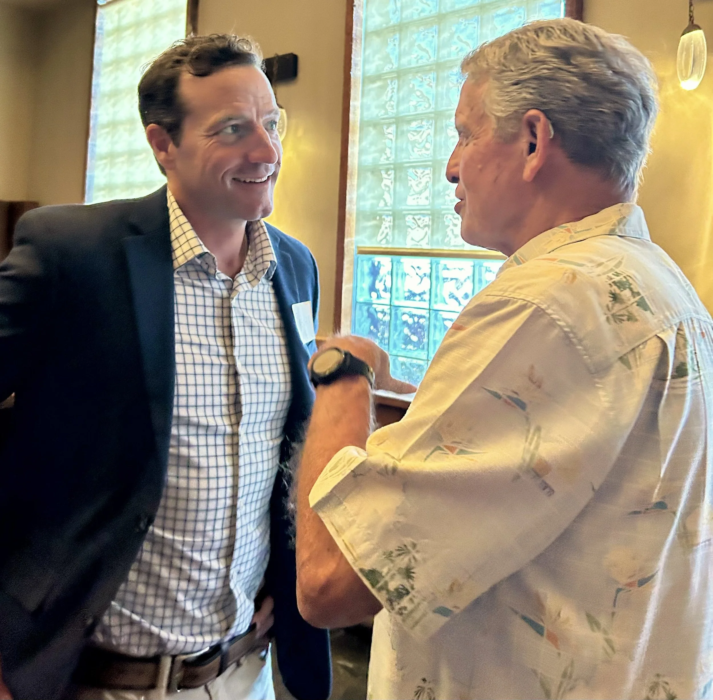 Brendan Moran having a friendly conversation indoors, one wearing a dark blazer and checkered shirt, the other in a floral short-sleeve shirt, standing near a window.
