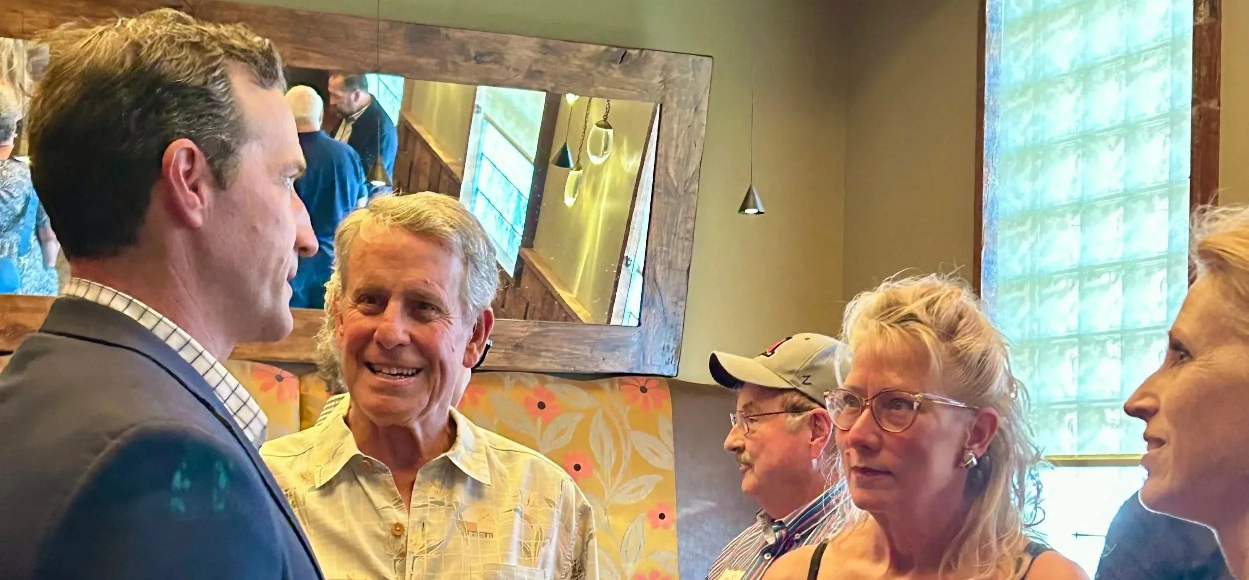 Five adults engaged in conversation inside a room with a large mirror, floral wallpaper, and hanging lighting.