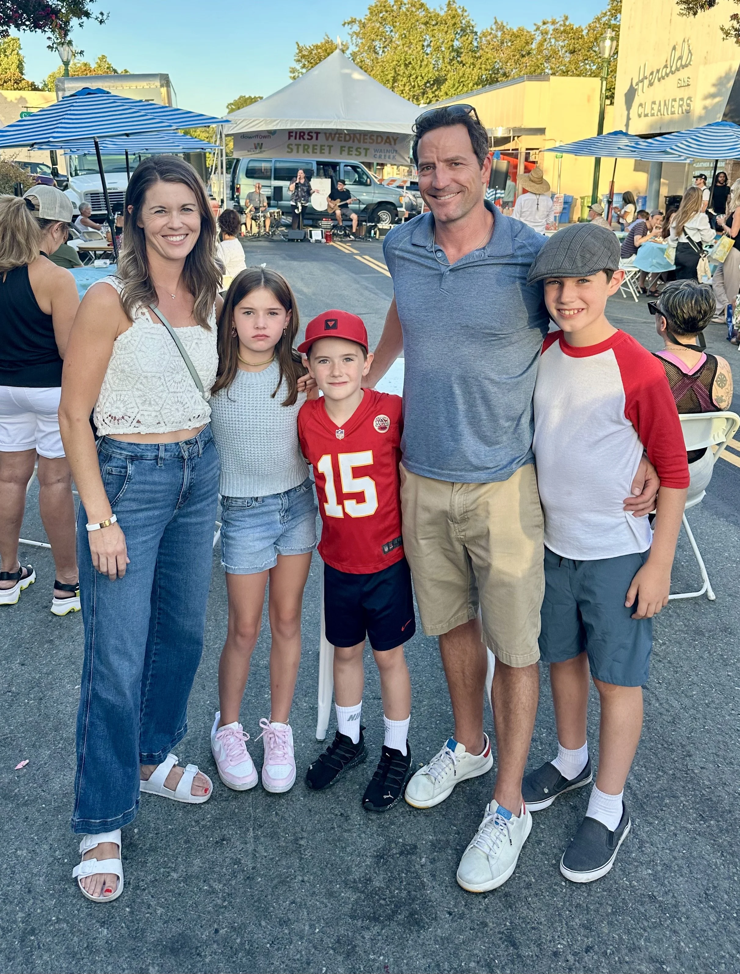Brendan Moran and family at an outdoor street festival with a stage and musicians in the background, people sitting at tables, and vendor tents. The family is smiling and posing for the photo.