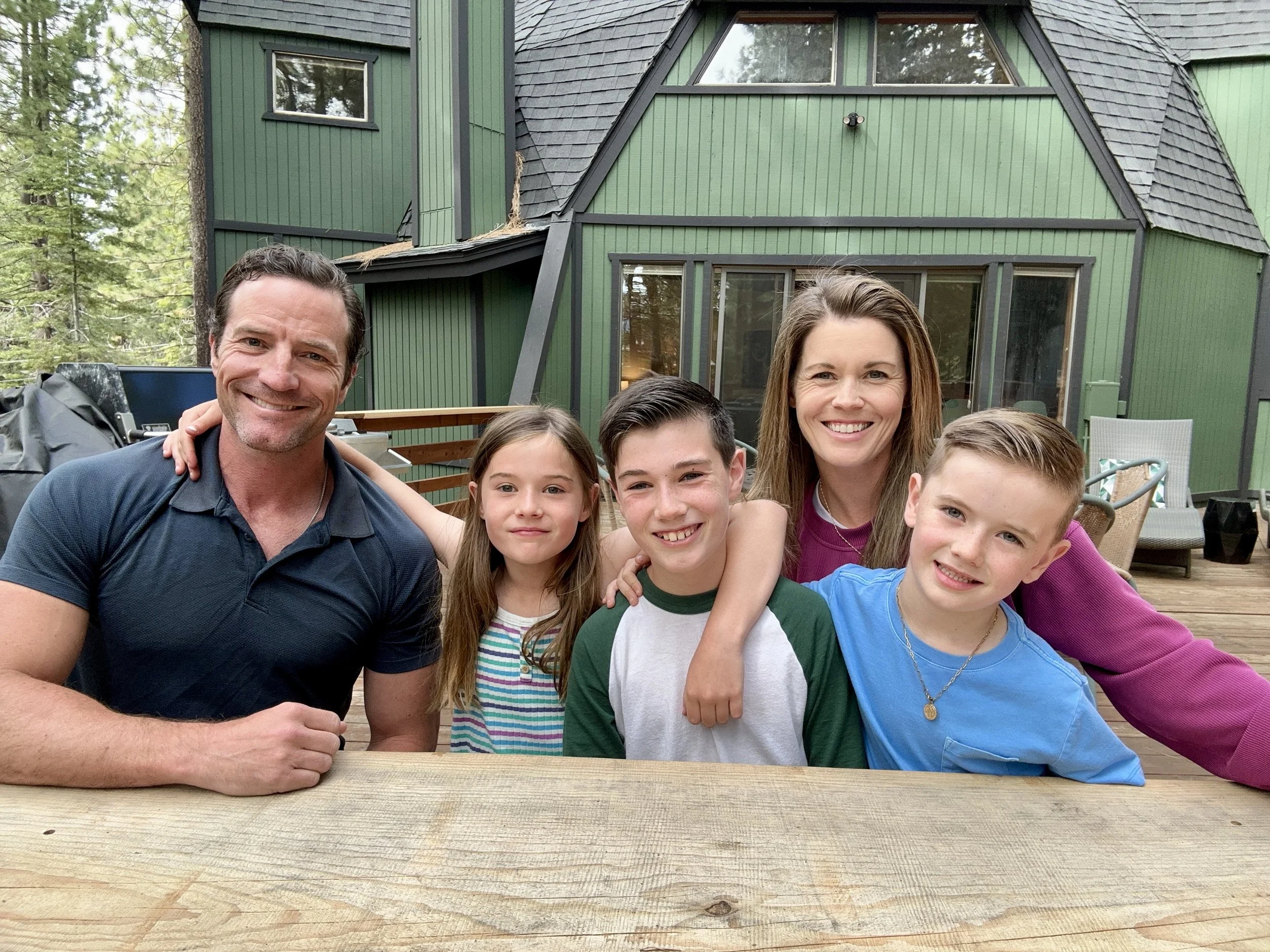 Family of Brendan Moran smiling and sitting outdoors on a wooden deck in front of a green house with large windows, surrounded by trees.