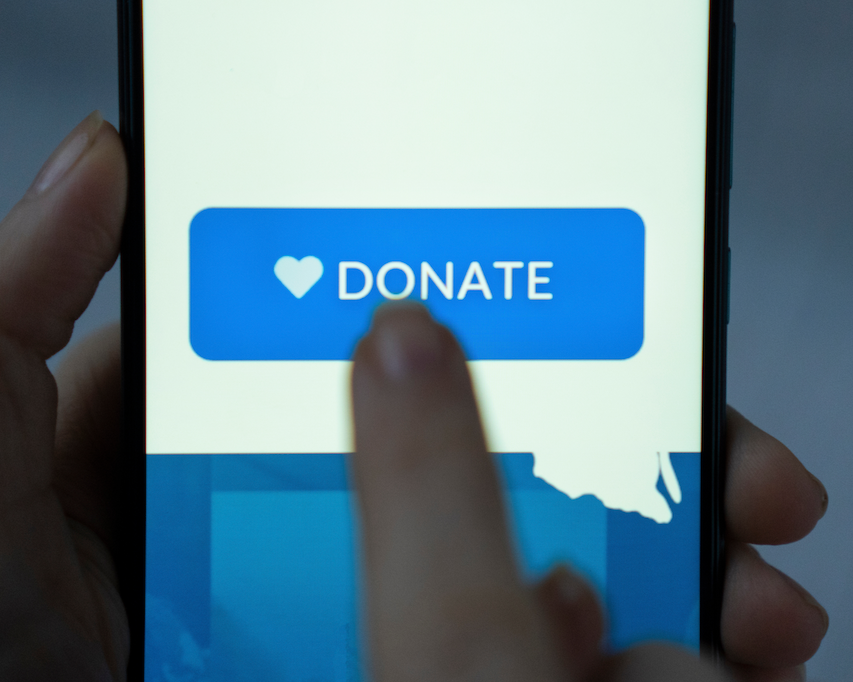 Donate to Brendan Moran. Close-up of a person tapping a smartphone screen with a blue 'DONATE' button featuring a heart icon on the screen.