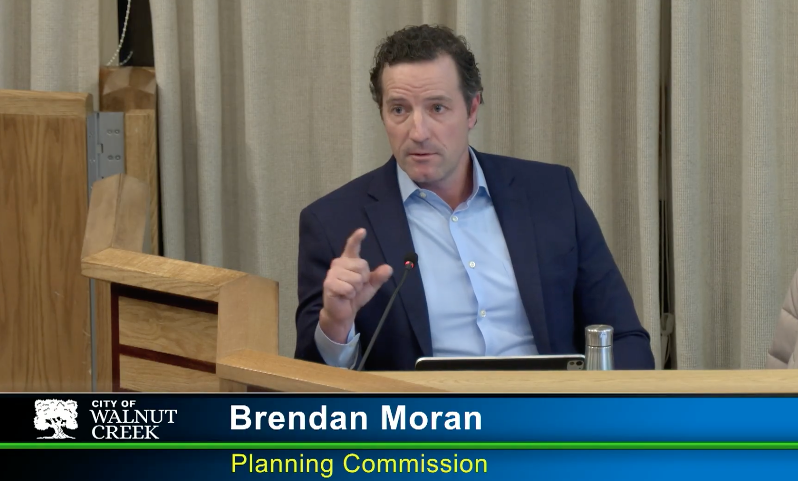 A man in a blue suit and light blue shirt is speaking at a meeting, sitting at a wooden table with a microphone, in front of beige curtains. The lower part of the image shows a banner with the text "City of Walnut Creek" and "Brendan Moran, Planning Commission."