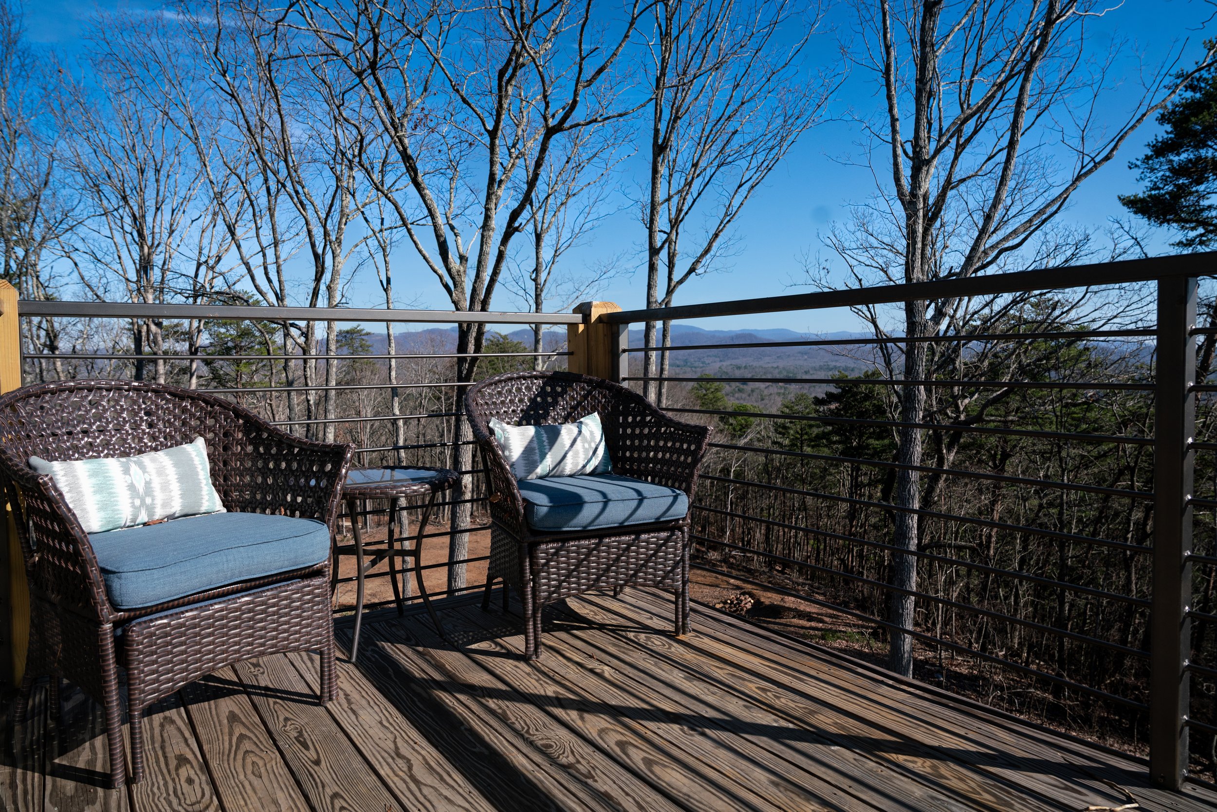 An outdoor wooden deck with two wicker chairs with blue cushions and pillows, a small matching table, overlooking a wooded landscape with leafless trees and distant mountains under a clear blue sky.