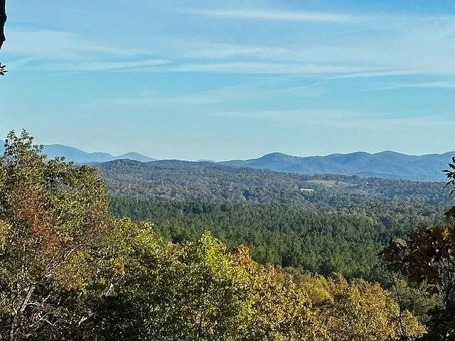View of rolling hills and mountains in the distance with a forested landscape in the foreground under a partly cloudy sky.