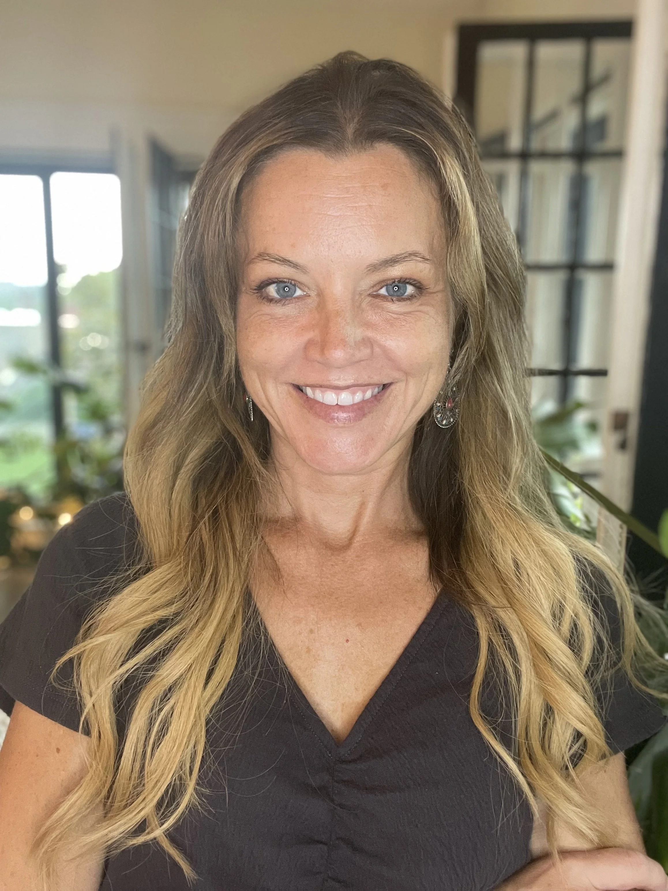 A woman with long, wavy blonde hair, blue eyes, wearing a black top, smiling, standing indoors with a window and plants in the background.