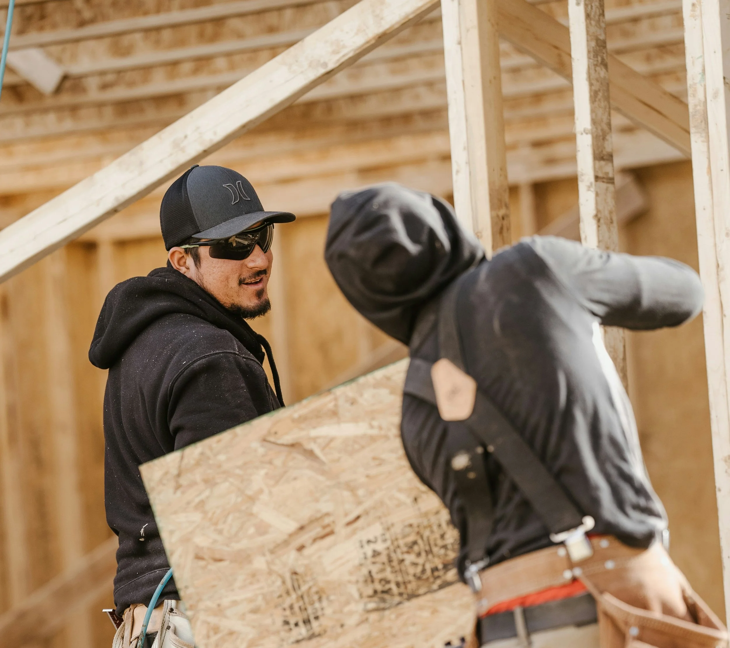 Two construction workers working inside a building under construction, with one man holding a piece of plywood and the other installing framing.