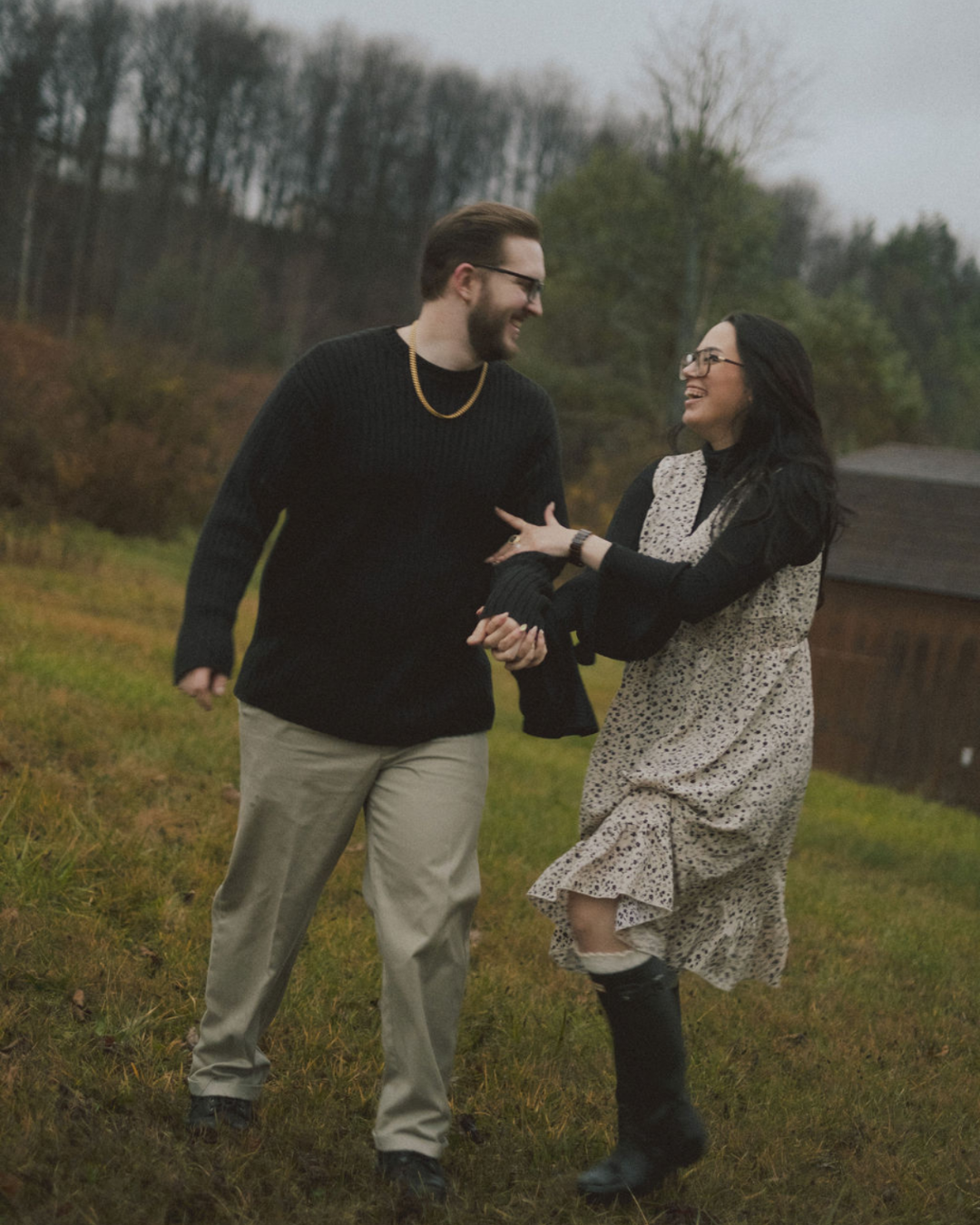 A man and woman outdoors on a grassy field, smiling and holding hands, with the woman playfully tugging on the man's arm. The background shows trees and a cloudy sky.