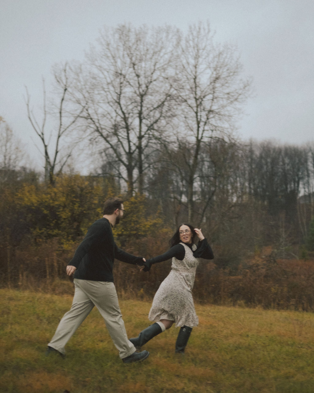 A man and woman holding hands and running outdoors on a fall day, with leafless trees and cloudy sky in the background.