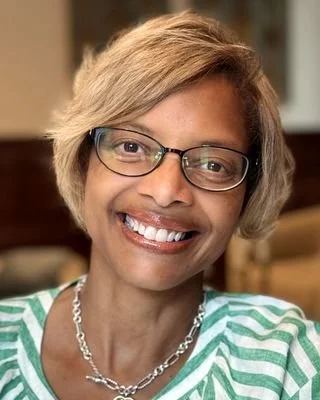 Smiling woman with short blonde hair, glasses, and a striped green and white top, wearing a silver chain necklace.
