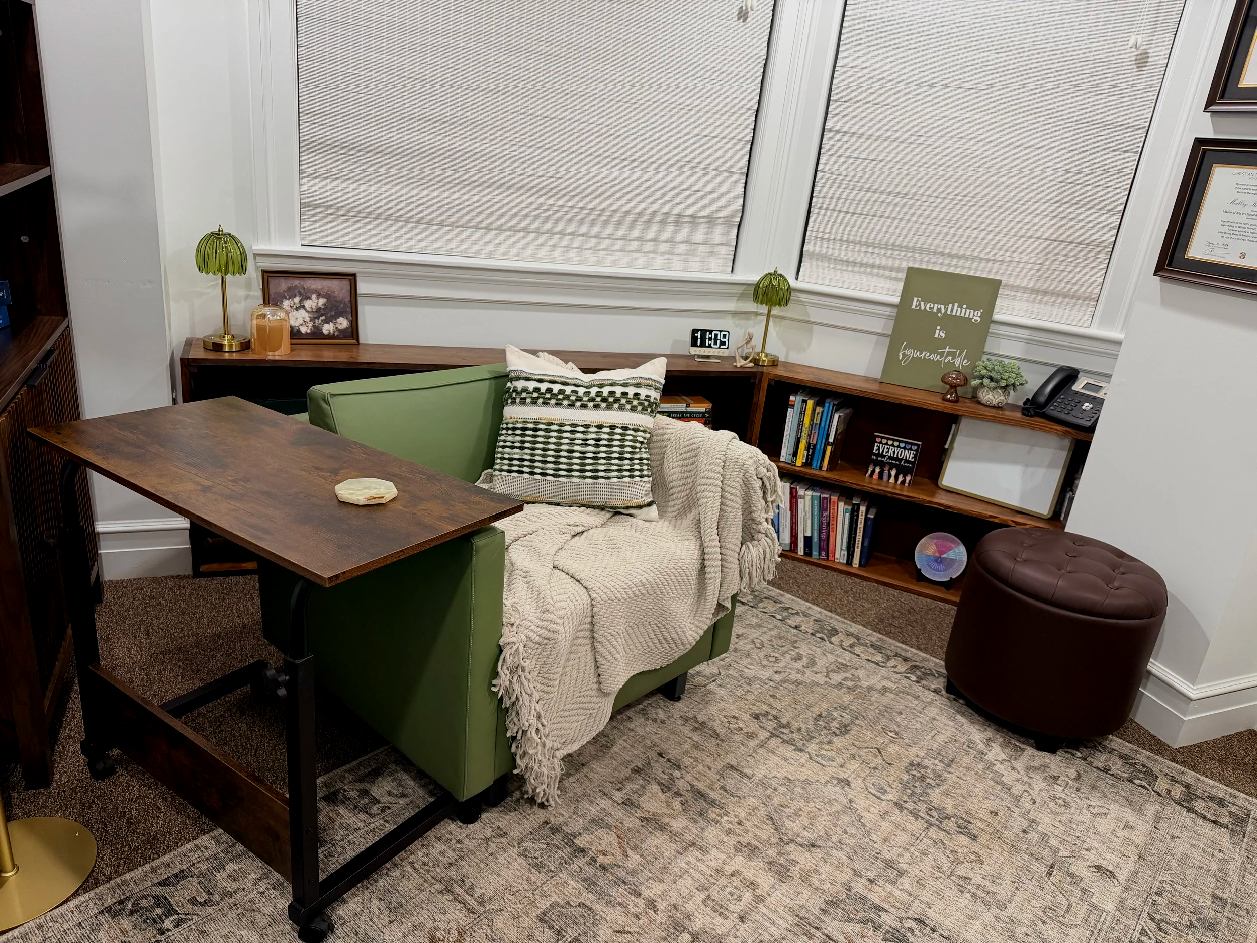 A cozy home office with a green couch covered with a beige blanket and decorative pillows, a wooden desk attached to the couch, a brown ottoman, and a wooden bookshelf with books, plants, and decorative items. The room has white walls, large windows with blinds, a clock, and framed artwork.
