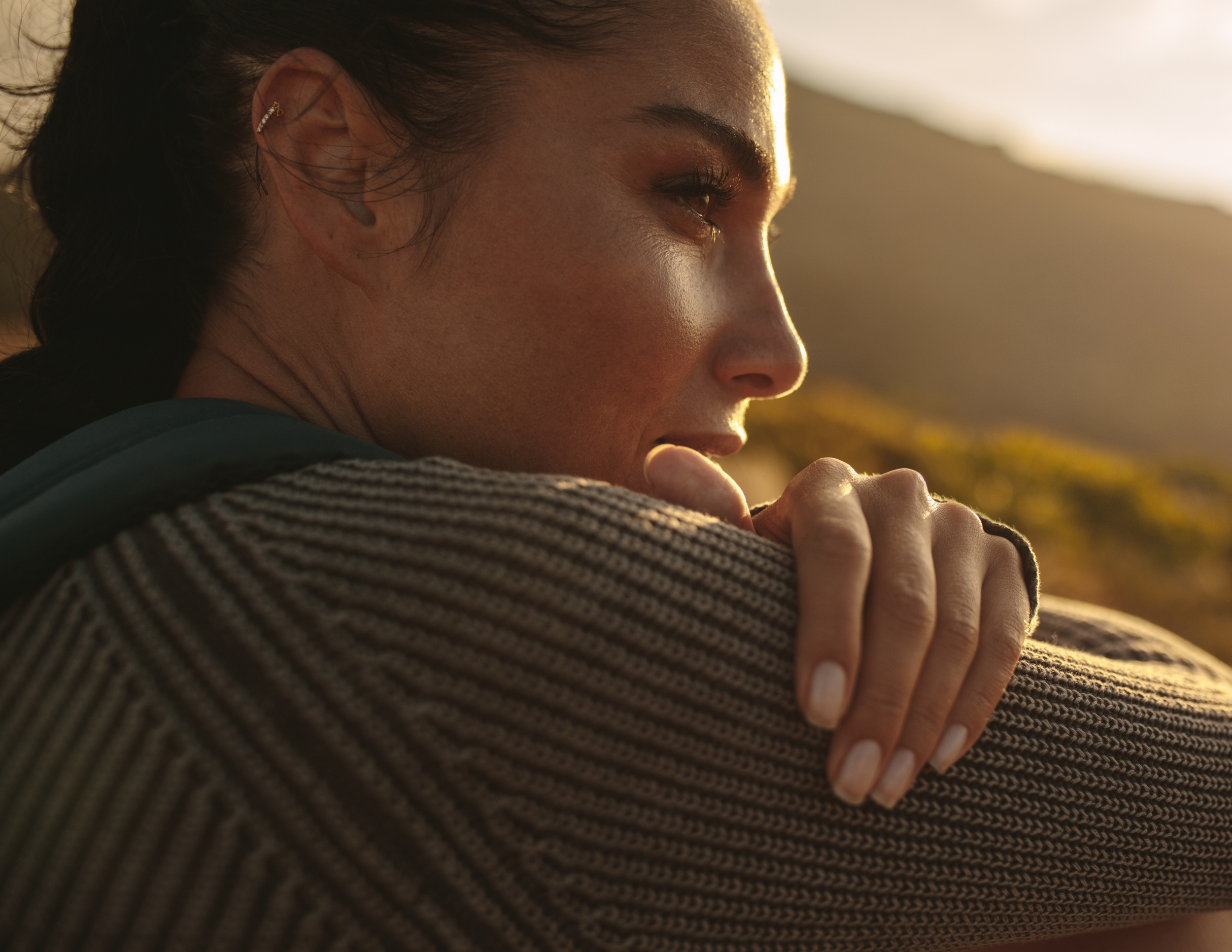 A woman with dark hair and earrings rests her chin on her folded arms, gazing thoughtfully into the distance during sunset.