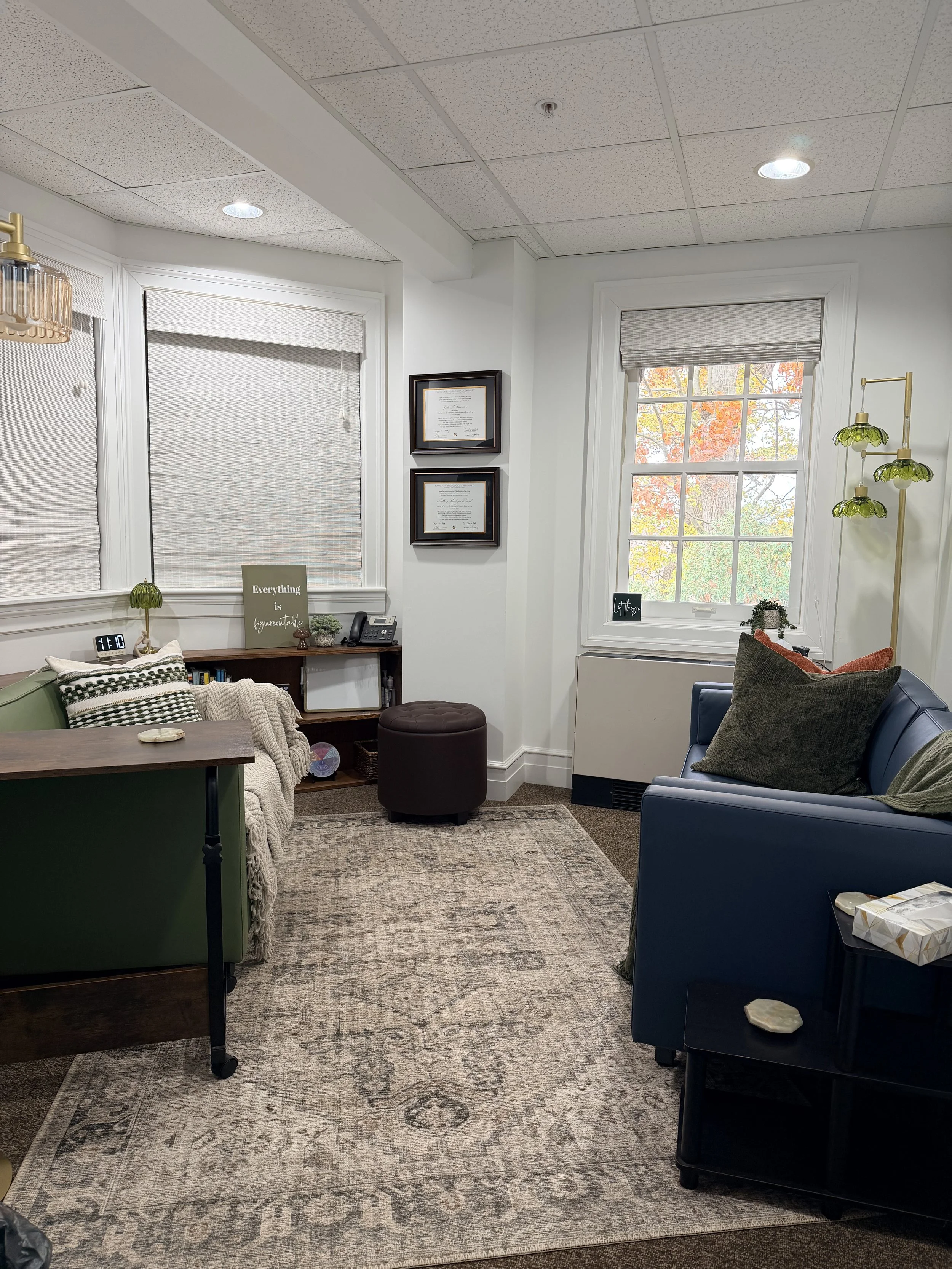 A cozy waiting room with two large windows, one with blinds closed and the other open showing fall foliage. The room has a green armchair, a dark blue sofa, and a small black side table with a tissue box. There is a beige patterned area rug, a brown ottoman, and a bookshelf with decorative items. The wall has framed certificates and a sign that reads 'Everything is figureoutable.' A floor lamp with green glass shades and a wooden ceiling light are also visible.