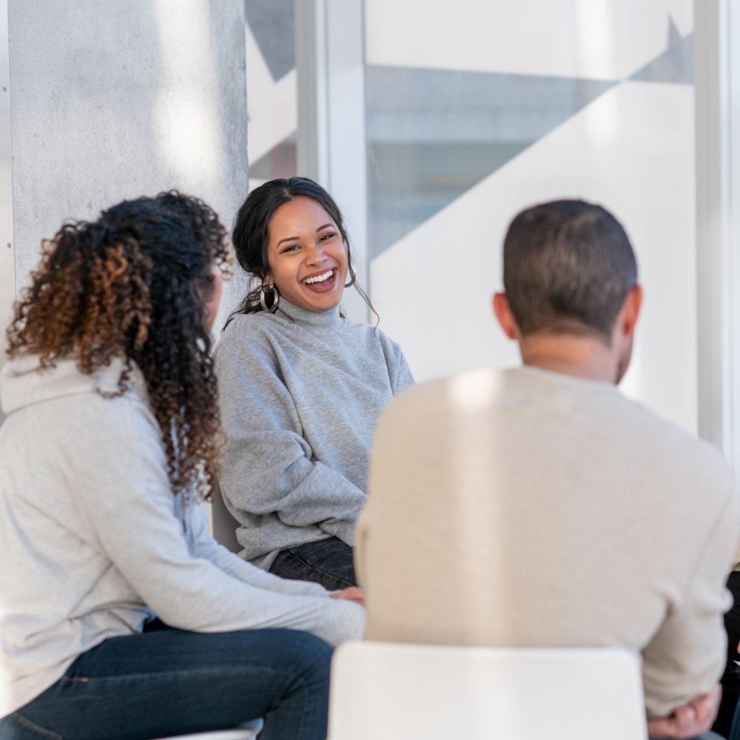 Three people sitting and talking in a modern, well-lit space, with a woman smiling and engaging in conversation.