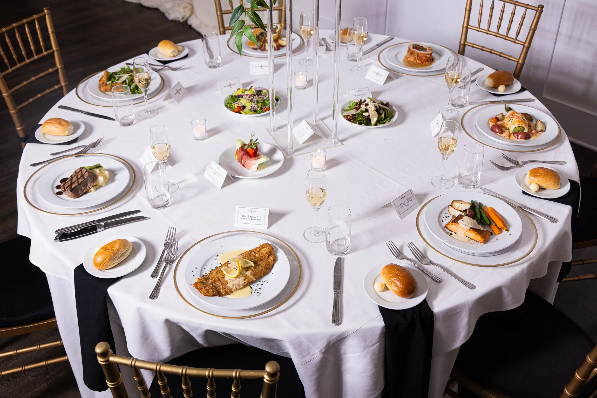 A round dining table set for a meal with a white tablecloth, gold trim plates, silverware, glasses of white wine or champagne, bread rolls, and small plates with various appetizers. Small candles and a centerpiece with green foliage and a tall glass vase add decoration to the table in a formal setting.