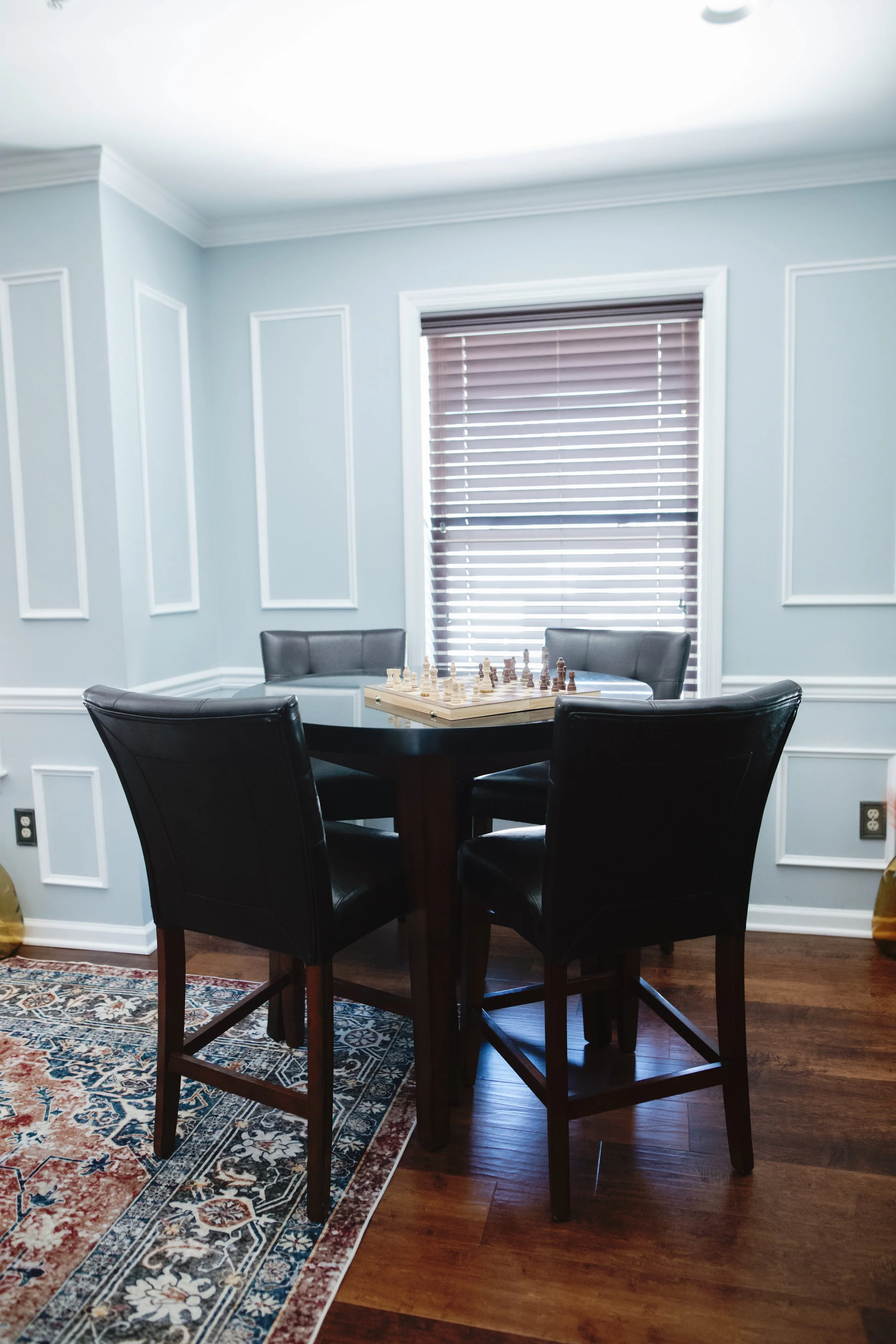 A dining area with a round black table, four black leather chairs, light blue walls with white trim, a window with closed blinds, and a chess set on the table.