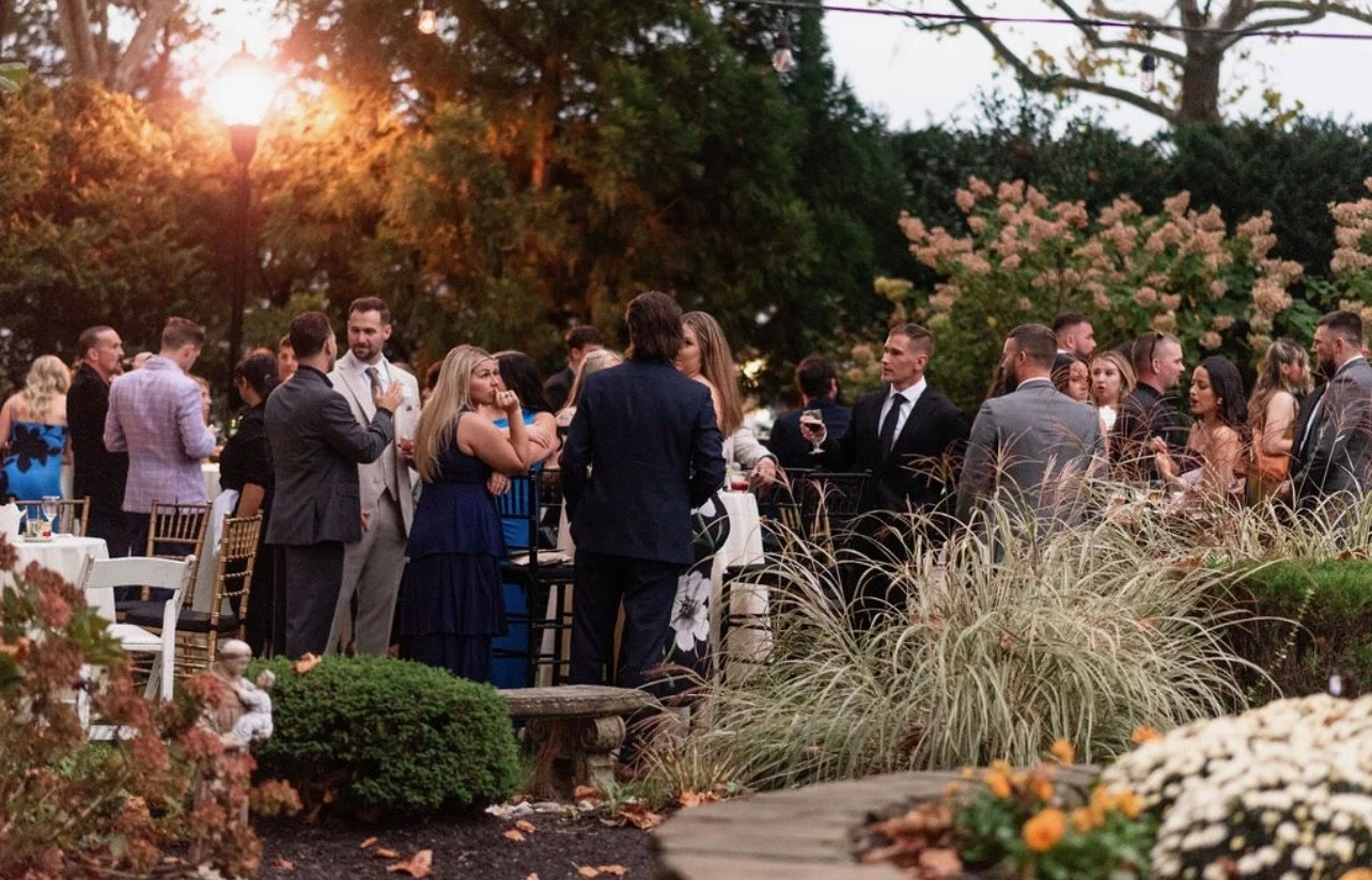 People at an outdoor social gathering or party in a garden during the early evening, with trees and flowering bushes in the background.