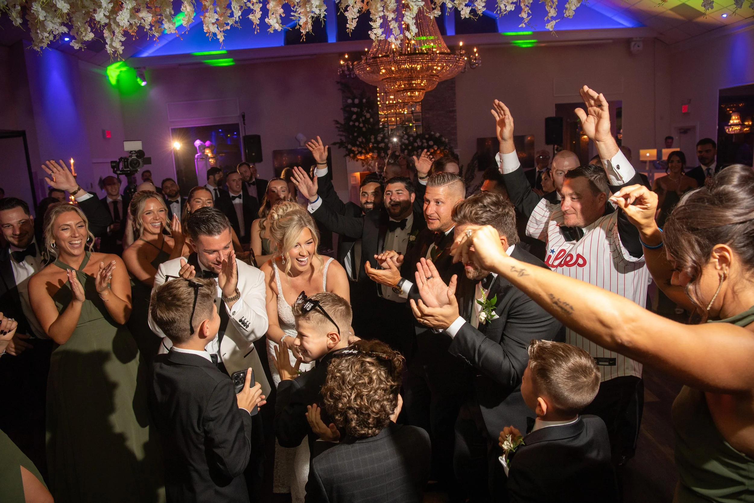 A wedding celebration with guests dancing and cheering in a decorated banquet hall, with a chandelier and floral arrangements.