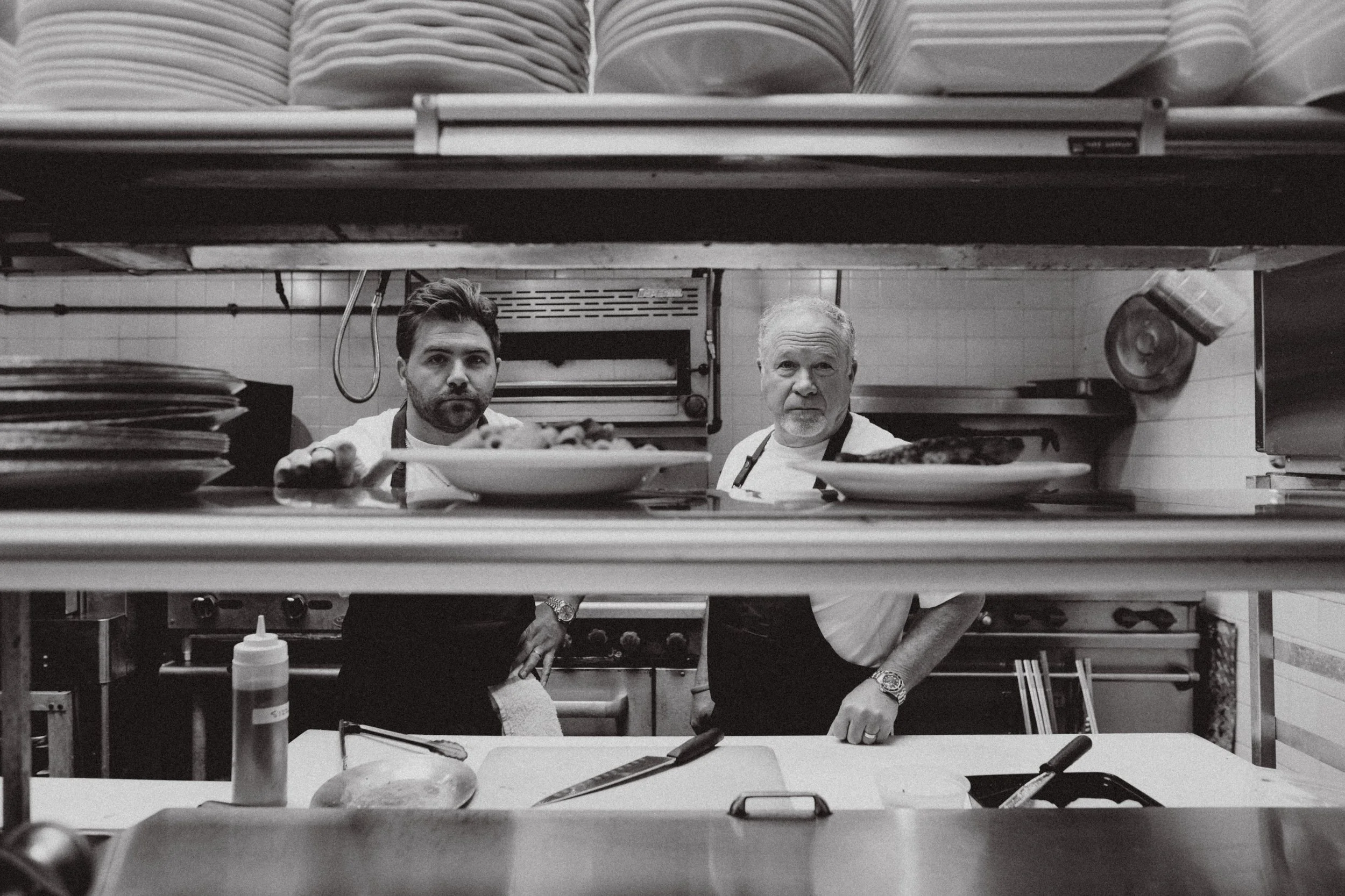 Two male chefs in a professional kitchen, viewed through a window in the kitchen wall, with plates of food in front of them and cooking equipment behind.