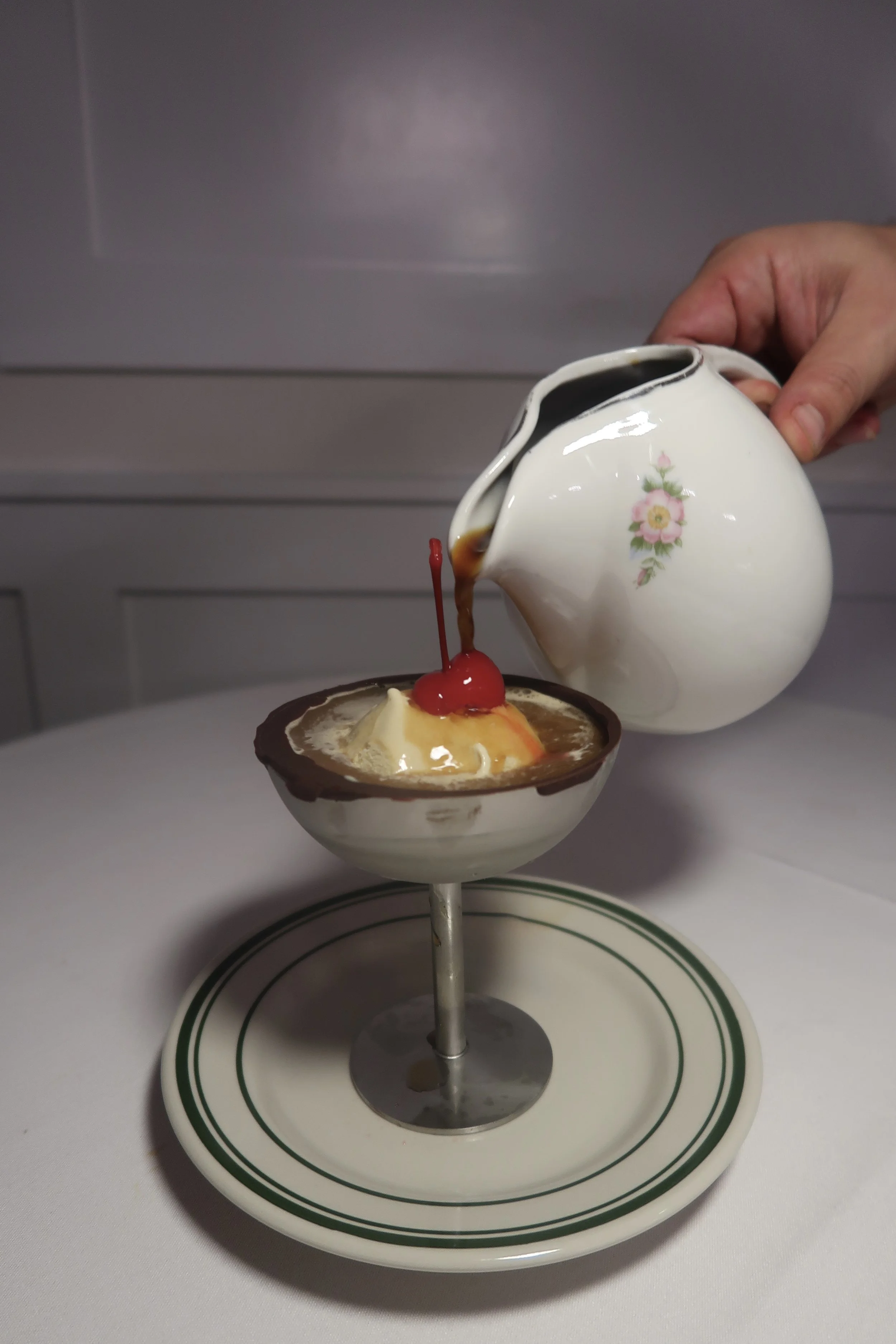 A hand pouring coffee from a floral teapot into a dessert cup topped with ice cream, a cherry, and caramel sauce.