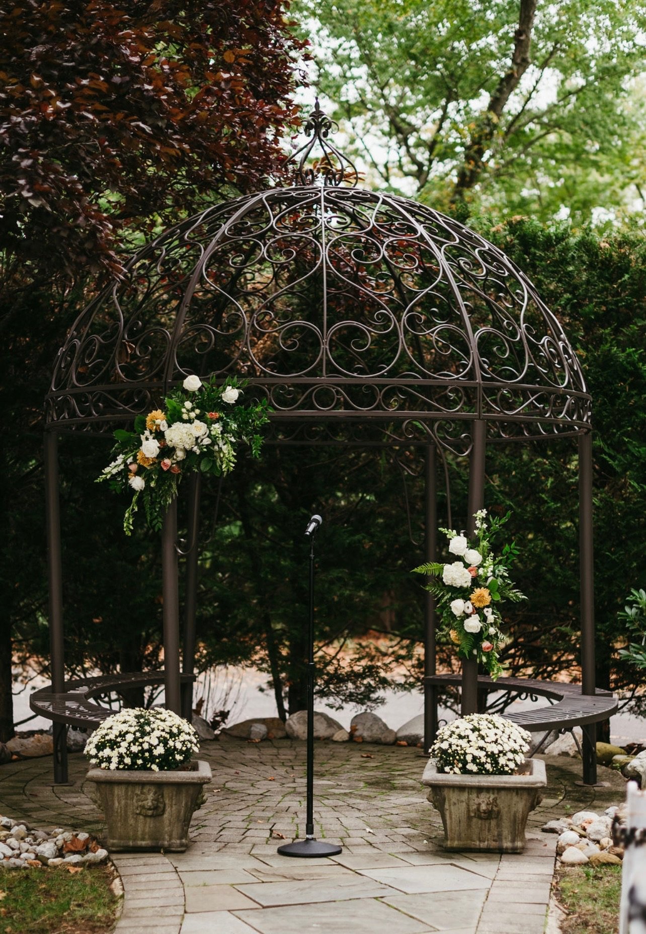 A decorative metal gazebo in a garden, decorated with flowers, with benches on either side and a microphone stand in front, surrounded by trees and plants.